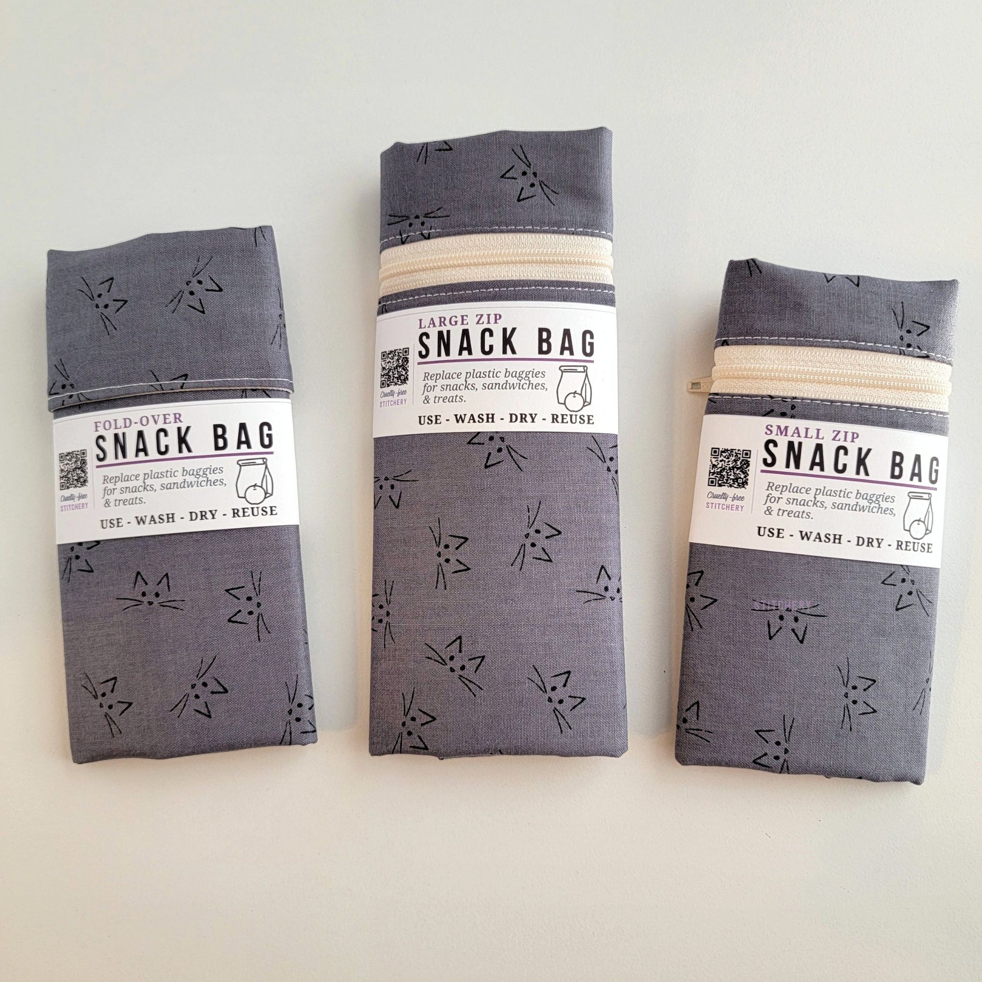 The Gray Cat Faces Snack Bags packaged up. They are folded to be the same width, around 3 inches, and secured with a paper tag wrapped around the middle that says Snack Bag - Replace plastic baggies for snacks, sandwiches, and treats, use, wash, dry, reuse, with an icon of a lunch bag and an apple.