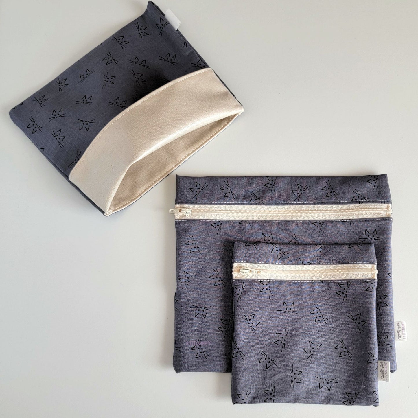 Set of three reusable snack bags with a gray cat faces print. Two have zippers, one large and one small, and one is a fold-over style. The fold-over one is unfolded to show the beige lining that matches the zippers. The print is a medium gray with very simply drawn cat faces - V shapes for ears, dots for eyes and nose, and whiskers.