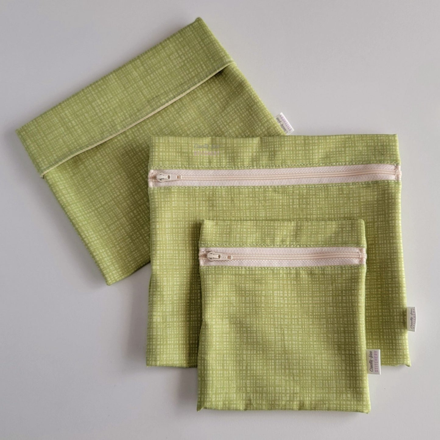 Set of three reusable snack bags with a light green textured print, shown with a foldover style bag closed and behind two different sizes of zipper styles. The print is a pale grass green with a cross-hatched texture.