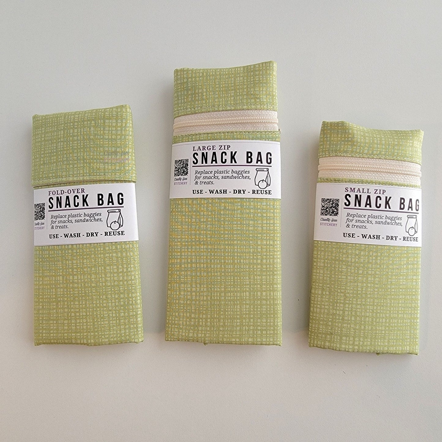 The Light Green Texture Snack Bags packaged up. They are folded to be the same width, around 3 inches, and secured with a paper tag wrapped around the middle that says Snack Bag - Replace plastic baggies for snacks, sandwiches, and treats, use, wash, dry, reuse, with an icon of a lunch bag and an apple. 
