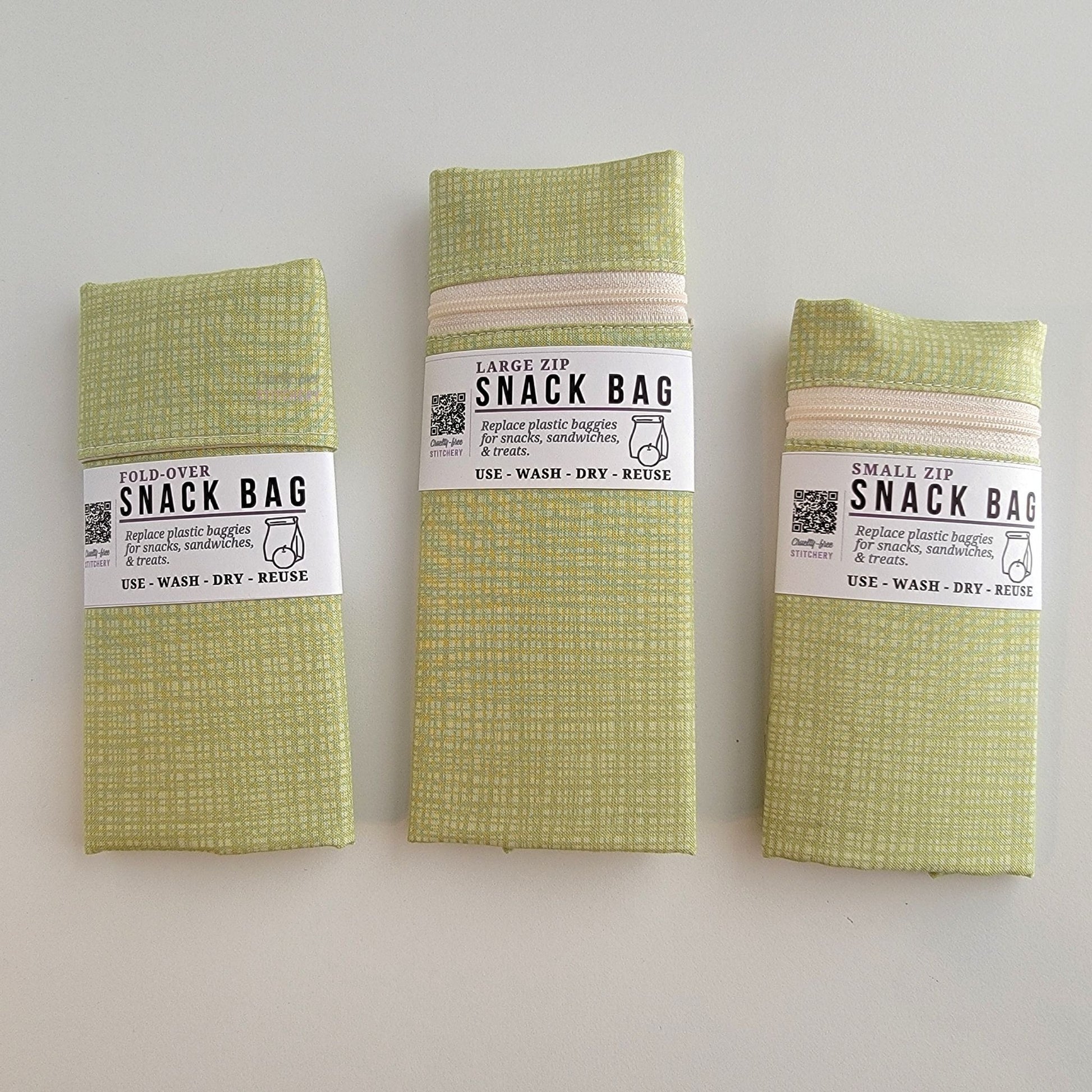 The Light Green Texture Snack Bags packaged up. They are folded to be the same width, around 3 inches, and secured with a paper tag wrapped around the middle that says Snack Bag - Replace plastic baggies for snacks, sandwiches, and treats, use, wash, dry, reuse, with an icon of a lunch bag and an apple. 