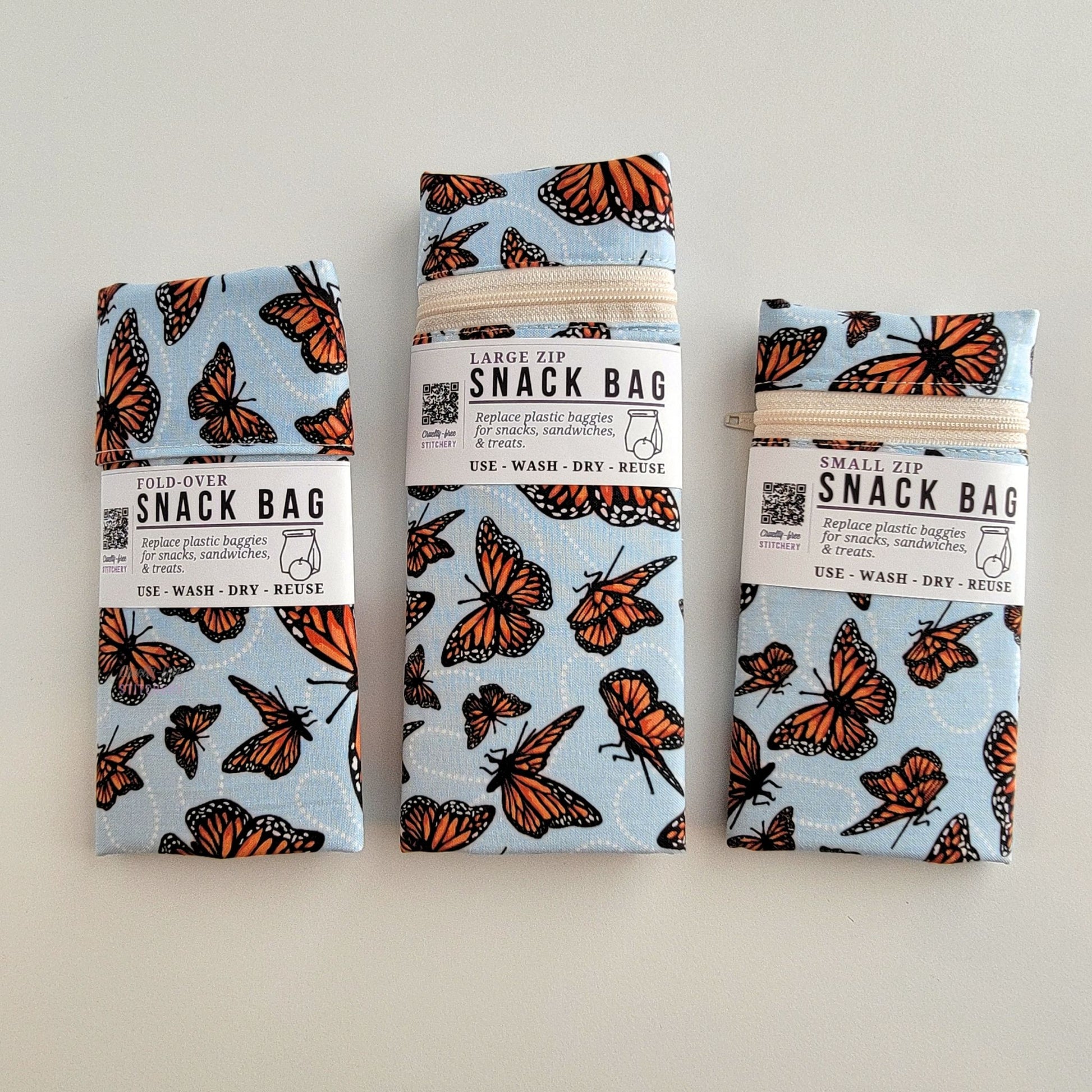 The Monarch Blue Snack Bags packaged up. They are folded to be the same width, around 3 inches, and secured with a paper tag wrapped around the middle that says Snack Bag - Replace plastic baggies for snacks, sandwiches, and treats, use, wash, dry, reuse, with an icon of a lunch bag and an apple. 