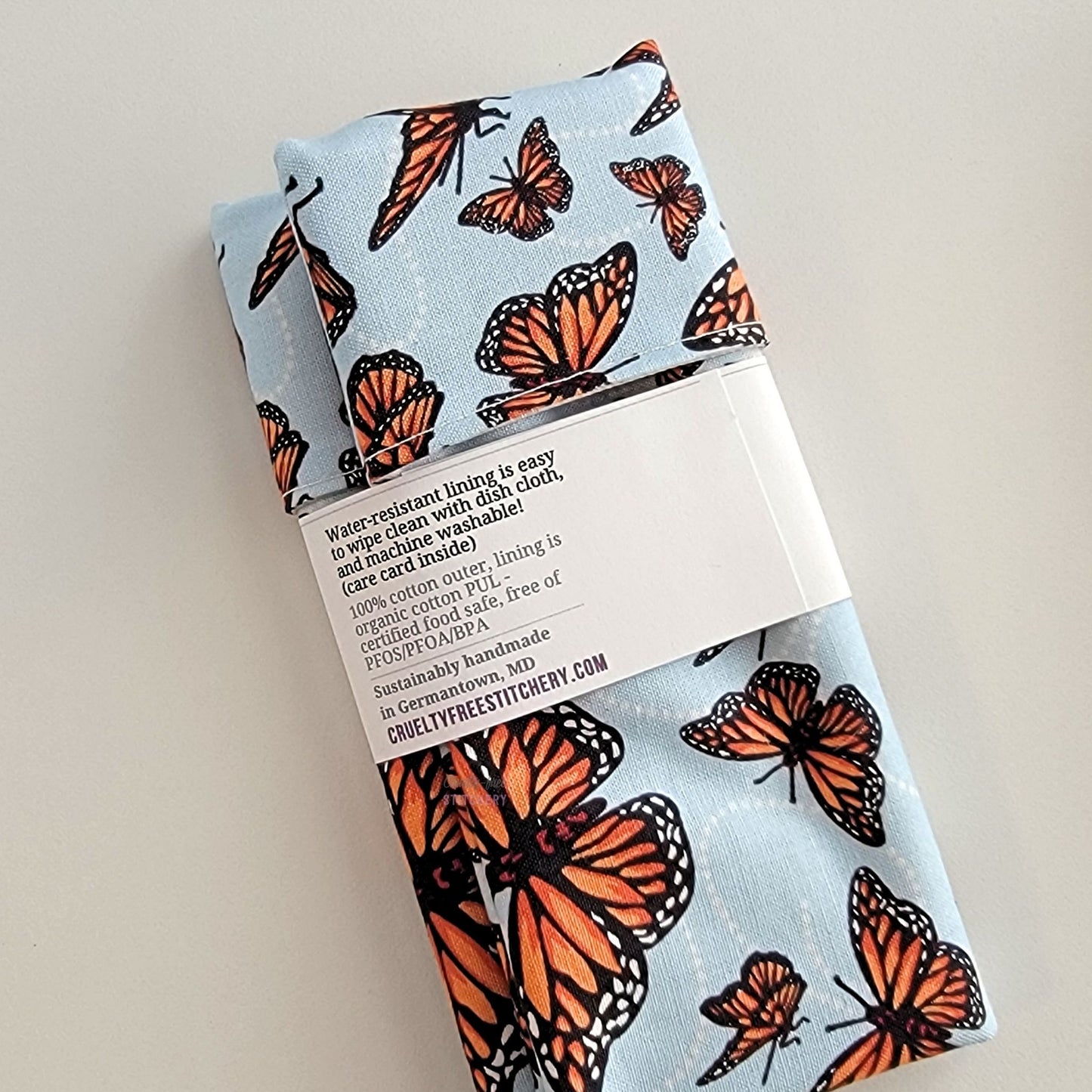 The back of the Monarch Blue Snack Bag packaging. It says "Water resistant lining is easy to wipe clean with dish cloth, and machine washable. Care card inside. 100% cotton outer, lining is organic cotton PUL, cerfitied food-safe, free of PFOS, PFOA, BPA. Sustainable handmade in Germantown, MD" and our website url.