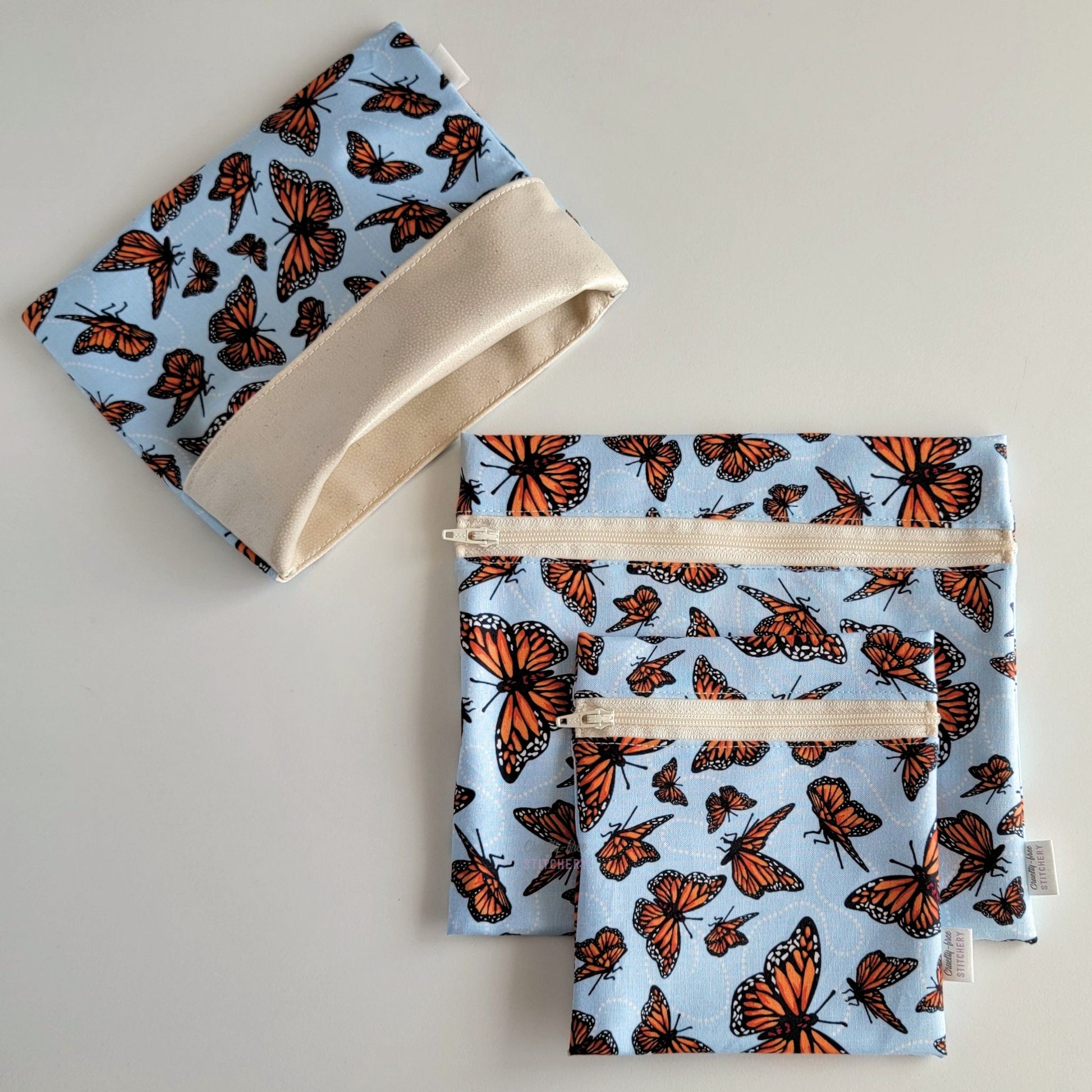 Set of three reusable snack bags with a Monarch butterfly pattern. Two have zippers, one large and one small, and one is a fold-over style. The fold-over one is unfolded to show the beige lining that matches the zippers. The fabric print is a light sky blue with orange monarch butterflies in varying sizes, with little white dots trailing behind them as they fly.
