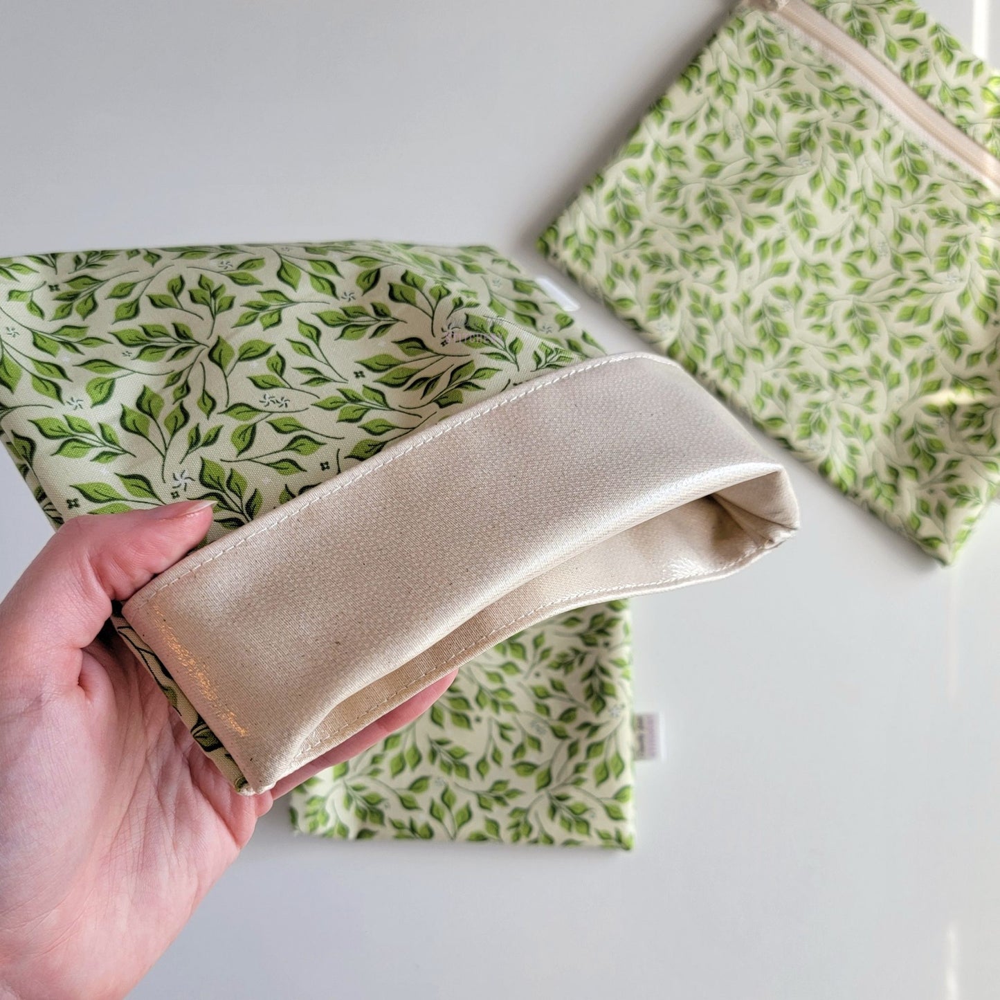 Hand holding an Olive Leaves Foldover Reusable Snack Bag open to show the lining inside, which is beige and is slightly shiny because it is water-resistant.
