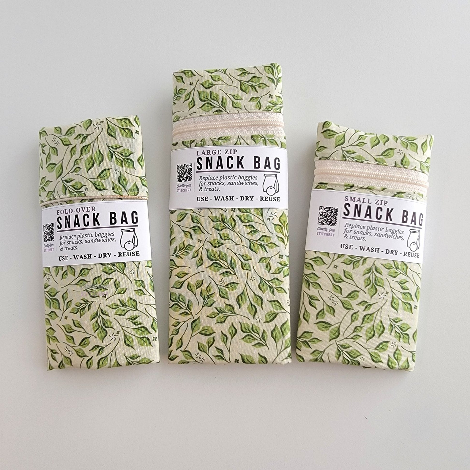 The Olive Leaves Snack Bags packaged up. They are folded to be the same width, around 3 inches, and secured with a paper tag wrapped around the middle that says Snack Bag - Replace plastic baggies for snacks, sandwiches, and treats, use, wash, dry, reuse, with an icon of a lunch bag and an apple. 