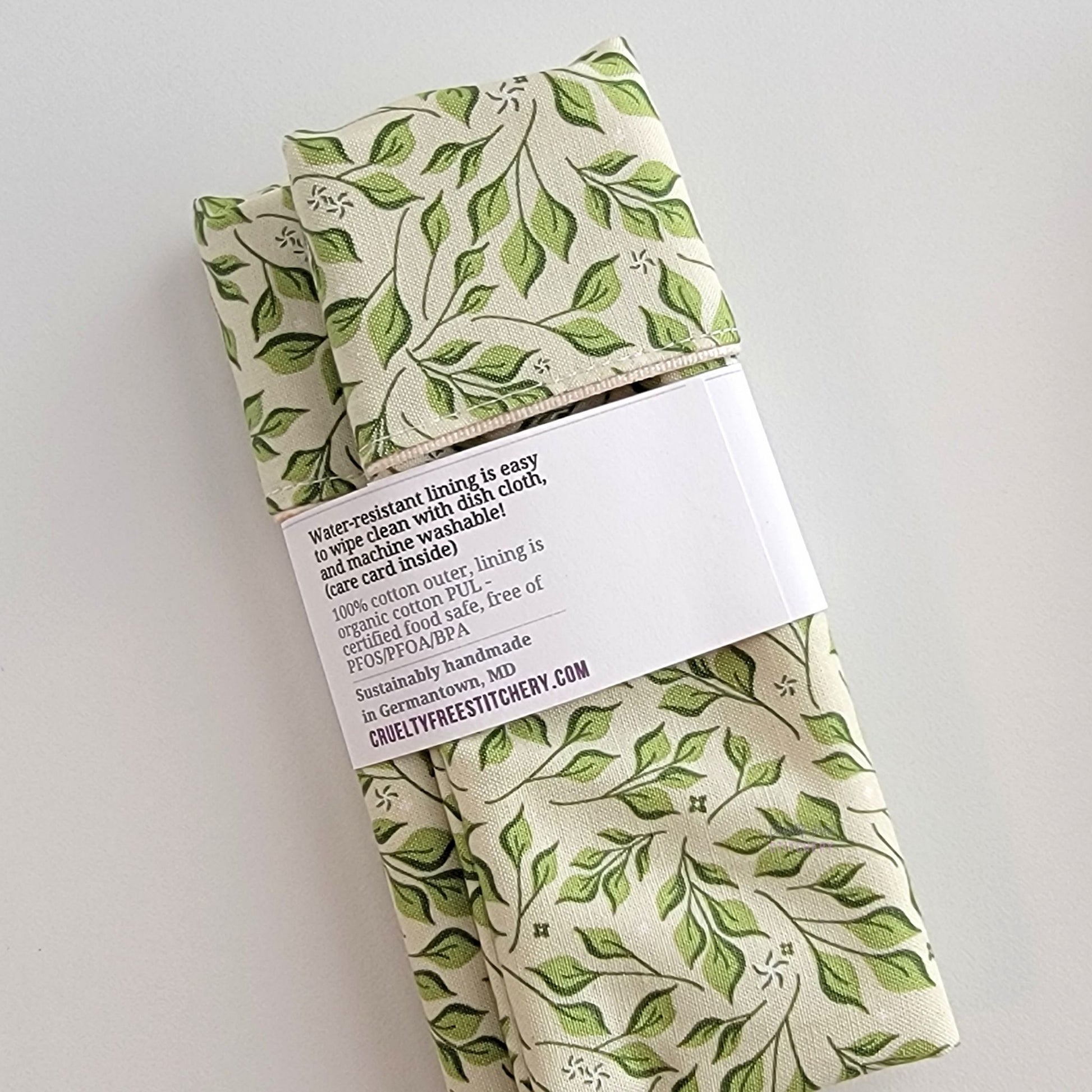 The back of the Olive Leaves Snack Bag packaging. It says "Water resistant lining is easy to wipe clean with dish cloth, and machine washable. Care card inside. 100% cotton outer, lining is organic cotton PUL, cerfitied food-safe, free of PFOS, PFOA, BPA. Sustainable handmade in Germantown, MD" and our website url.
