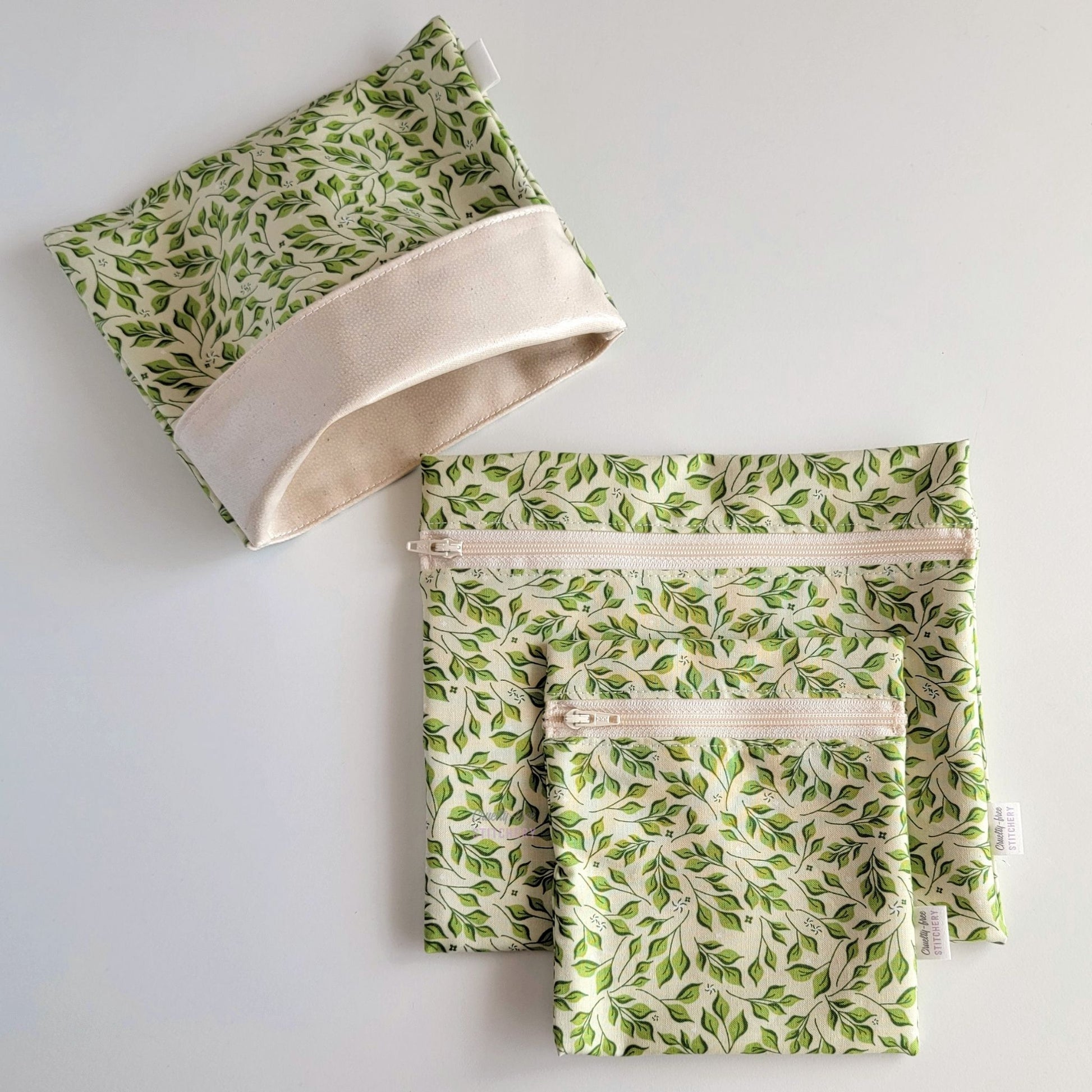 Set of three reusable snack bags with a light green leaves print. Two have zippers, one large and one small, and one is a fold-over style. The fold-over one is unfolded to show the beige lining that matches the zippers.