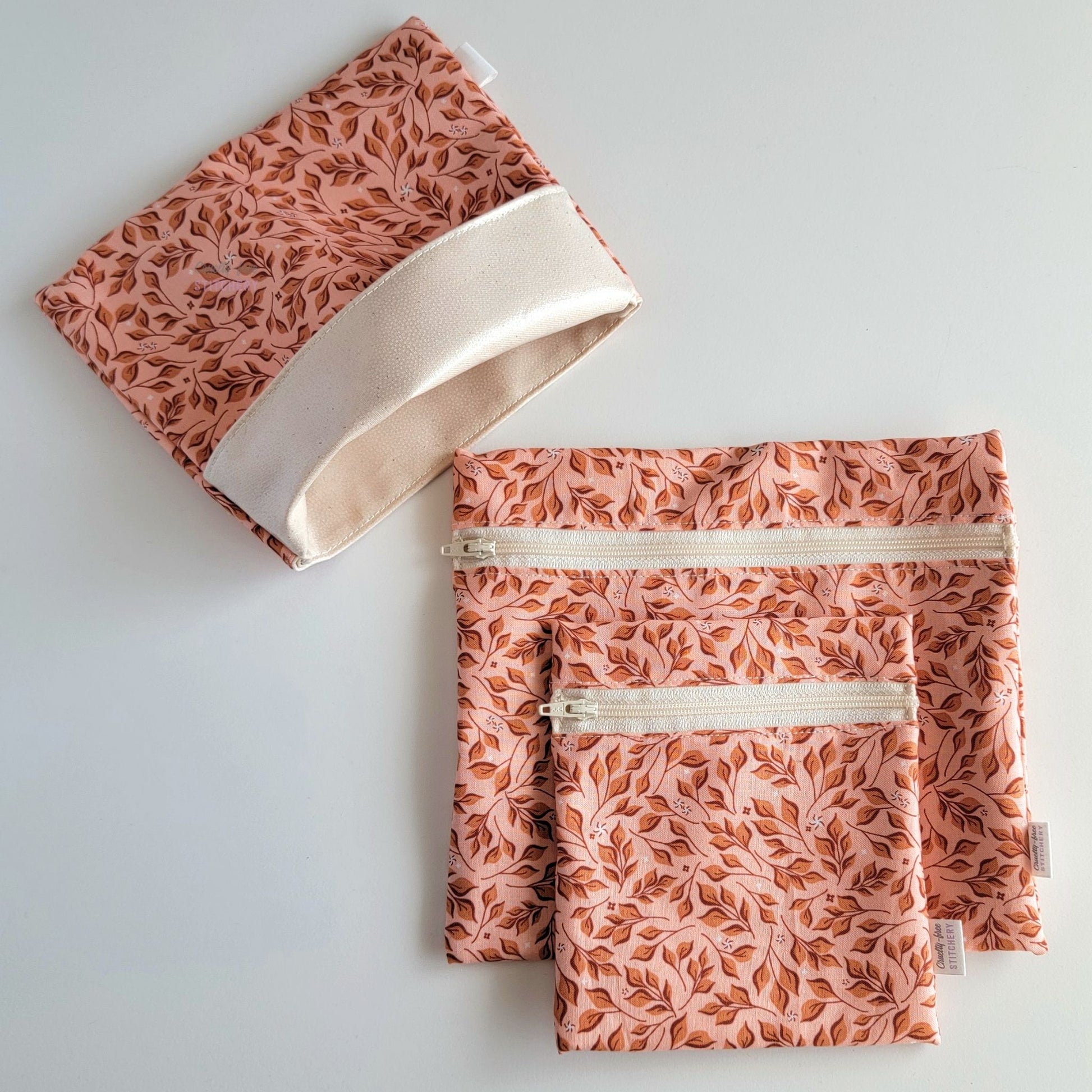 Set of three reusable snack bags with a pink leaves print. Two have zippers, one large and one small, and one is a fold-over style. The fold-over one is unfolded to show the beige lining that matches the zippers.
