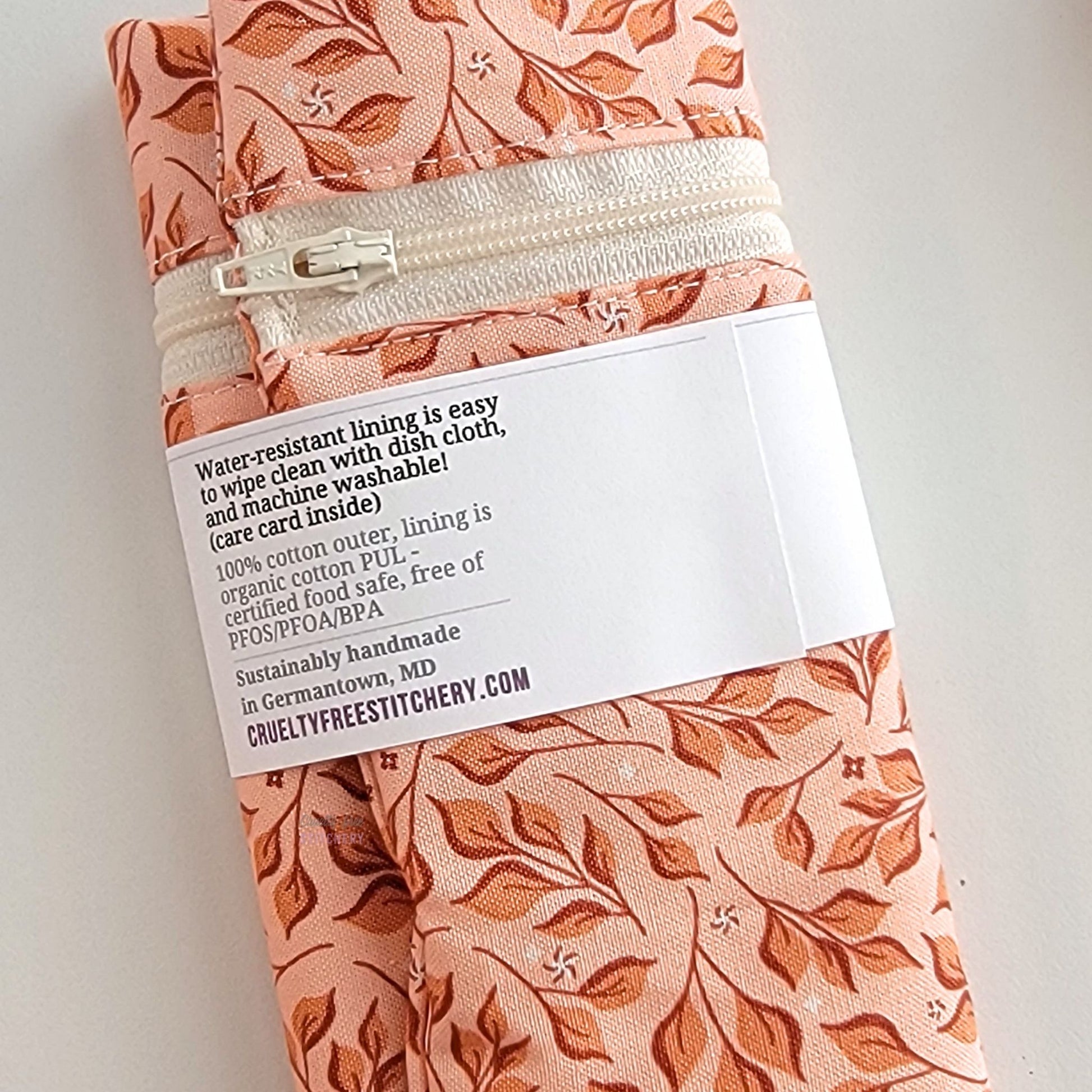 The back of the Pink Leaves Snack Bag packaging. It says "Water resistant lining is easy to wipe clean with dish cloth, and machine washable. Care card inside. 100% cotton outer, lining is organic cotton PUL, cerfitied food-safe, free of PFOS, PFOA, BPA. Sustainable handmade in Germantown, MD" and our website url.