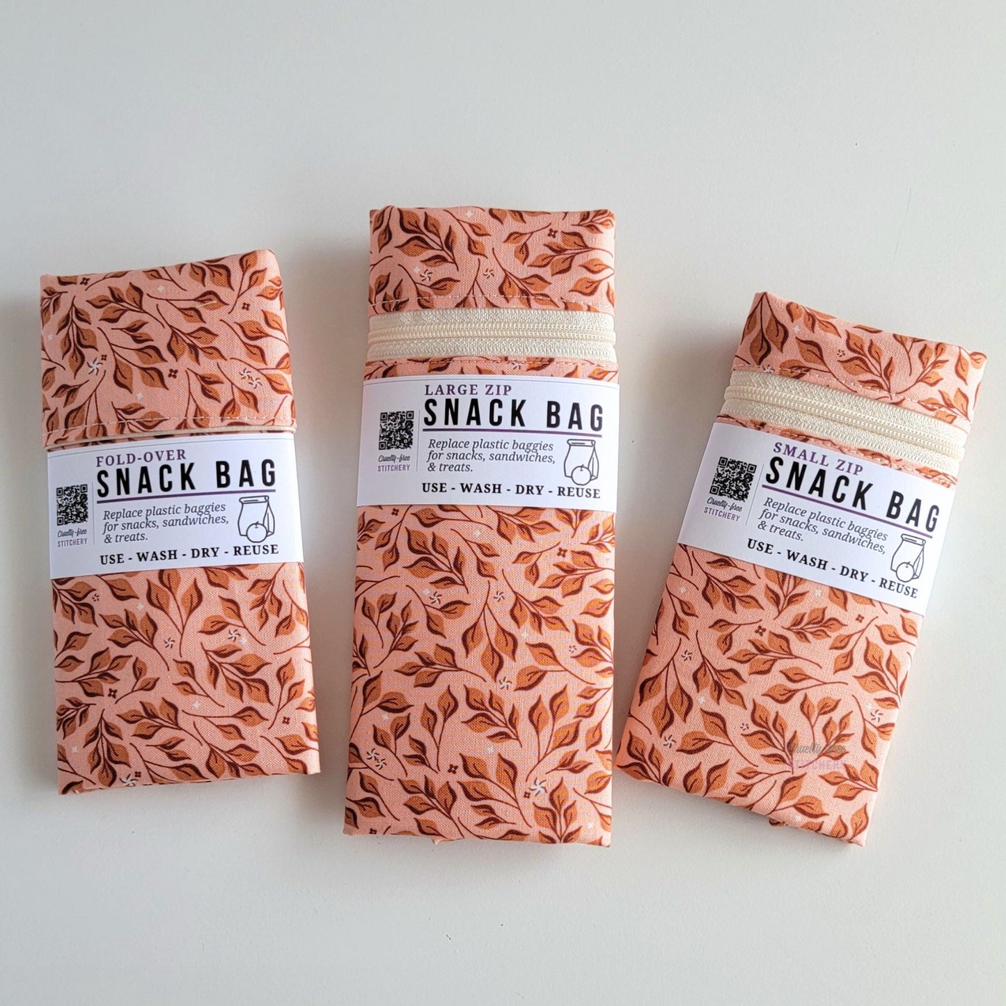The Pink Leaves Snack Bags packaged up. They are folded to be the same width, around 3 inches, and secured with a paper tag wrapped around the middle that says Snack Bag - Replace plastic baggies for snacks, sandwiches, and treats, use, wash, dry, reuse, with an icon of a lunch bag and an apple. 