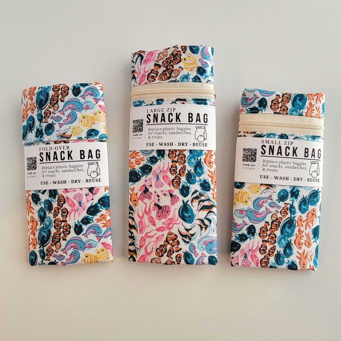 The Reef Snack Bags packaged up. They are folded to be the same width, around 3 inches, and secured with a paper tag wrapped around the middle that says Snack Bag - Replace plastic baggies for snacks, sandwiches, and treats, use, wash, dry, reuse, with an icon of a lunch bag and an apple. 