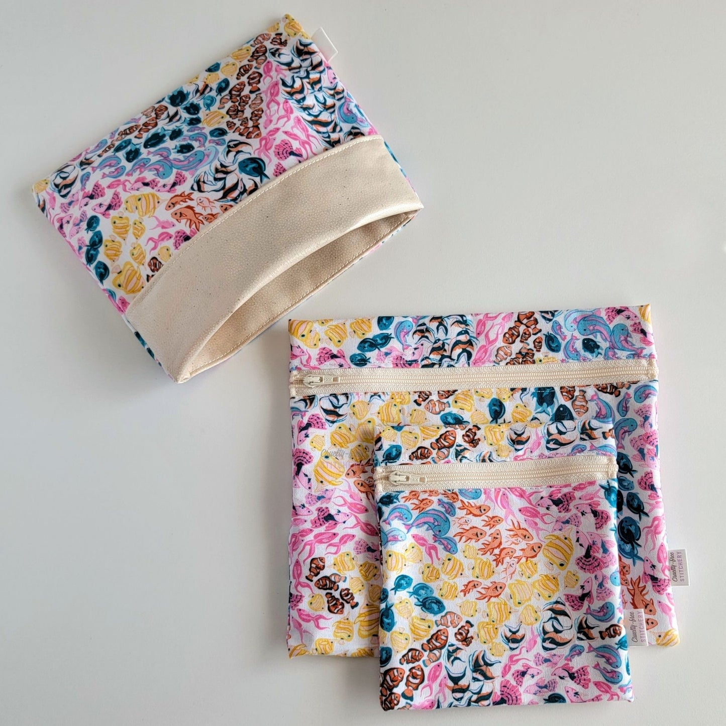 Set of three reusable snack bags with a bright fish print. Two have zippers, one large and one small, and one is a fold-over style. The fold-over one is unfolded to show the beige lining that matches the zippers. The print is a white background with swirls of small colorful tropical fish. Colors of yellow, pink, blue, and orange.
