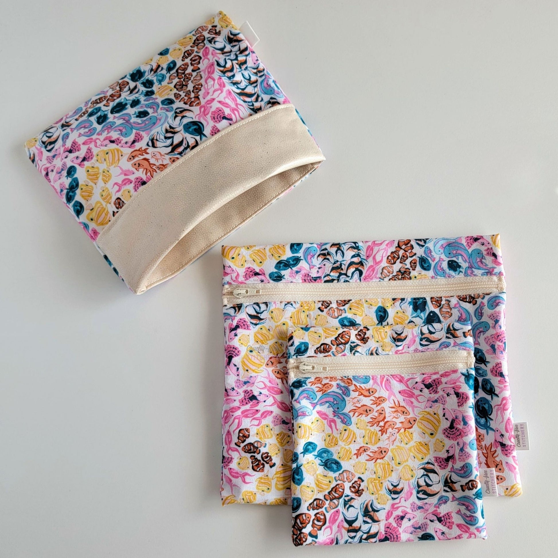 Set of three reusable snack bags with a bright fish print. Two have zippers, one large and one small, and one is a fold-over style. The fold-over one is unfolded to show the beige lining that matches the zippers. The print is a white background with swirls of small colorful tropical fish. Colors of yellow, pink, blue, and orange.

