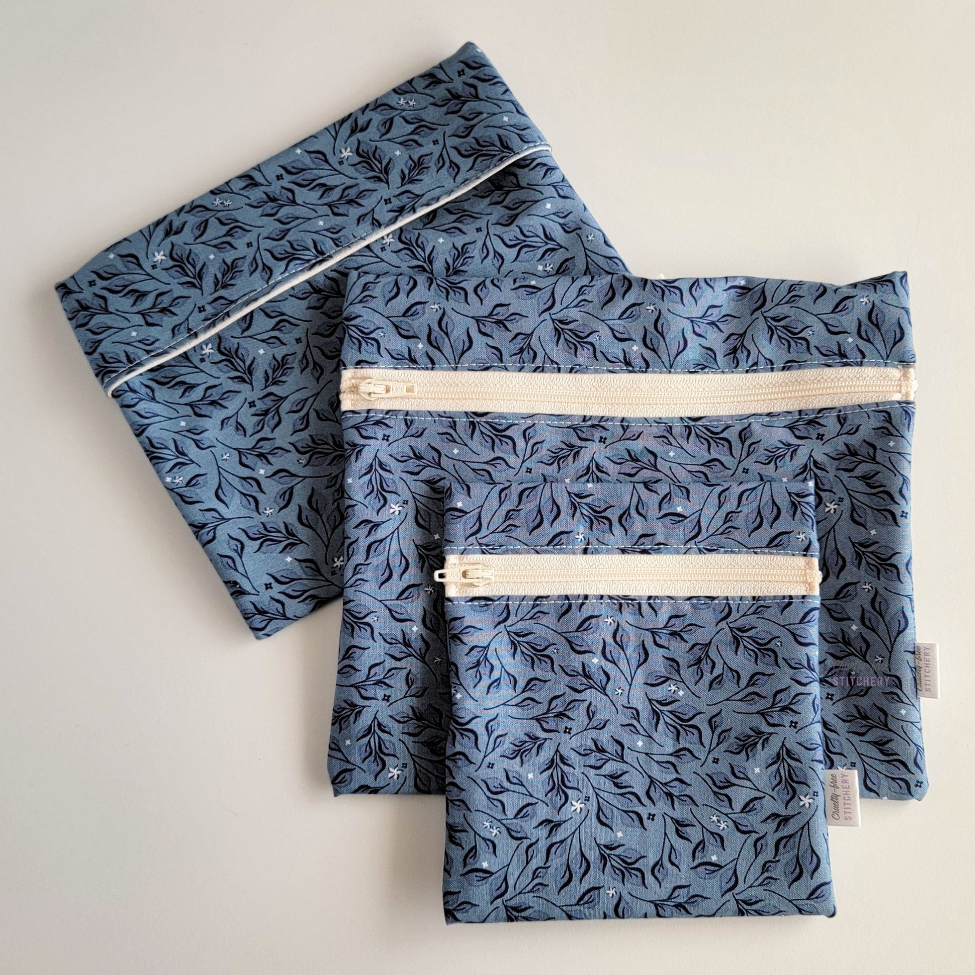 Set of three reusable snack bags with a dark blue leaves print, shown with the foldover style bag closed and behind the zipper styles. The print is a dark blue with darker navy leaves and stems.