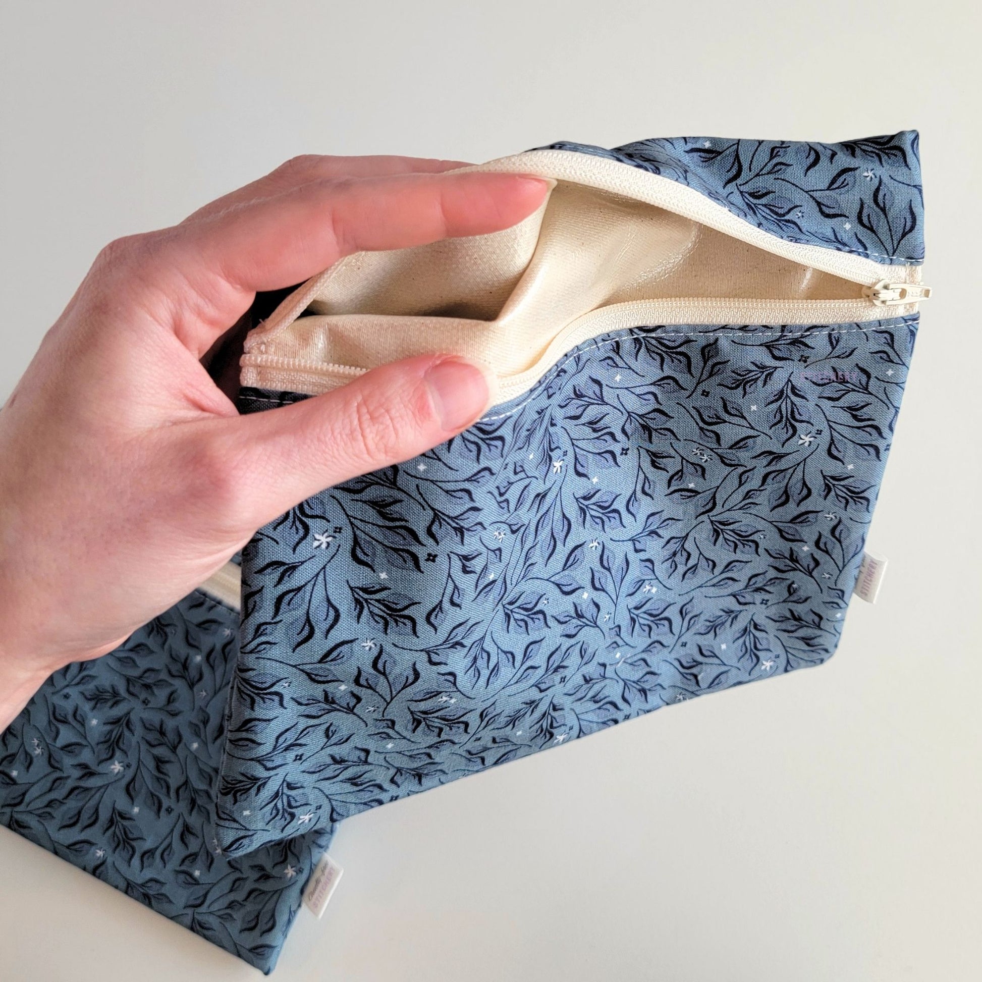 Hand holding a Stone Leaves Large Zip Reusable Snack Bag open to show the lining inside, which is beige and is slightly shiny because it is water-resistant.

