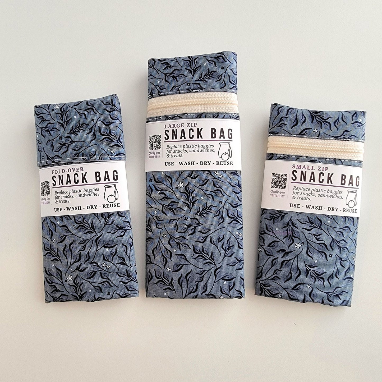 The Stone Leaves Snack Bags packaged up. They are folded to be the same width, around 3 inches, and secured with a paper tag wrapped around the middle that says Snack Bag - Replace plastic baggies for snacks, sandwiches, and treats, use, wash, dry, reuse, with an icon of a lunch bag and an apple. 