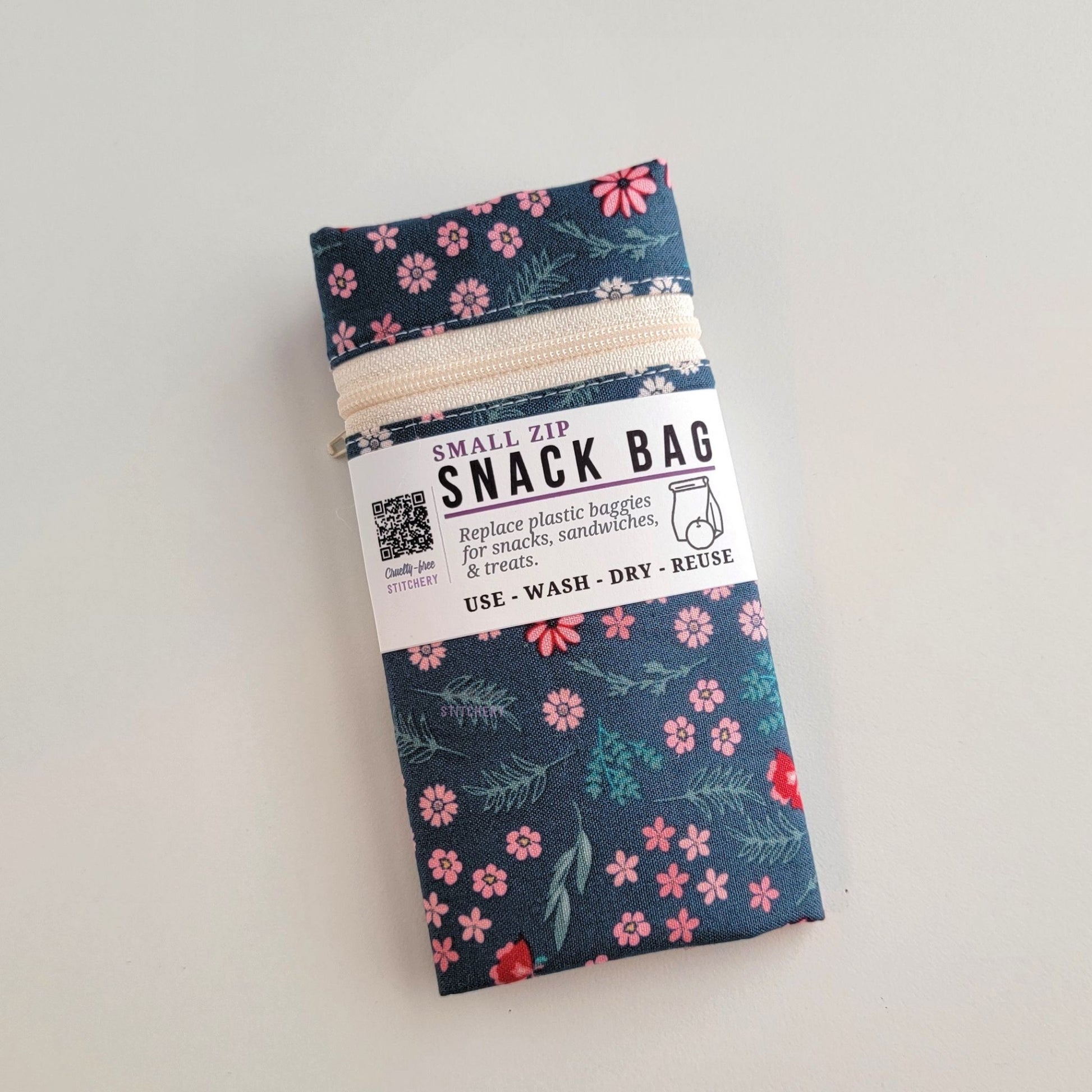 A Teal Pink Floral snack bag packaged up, it is folded to about 3 inches wide and wrapped with a paper band that says Snack Bag - Replace plastic baggies for snacks, sandwiches, and treats, use, wash, dry, reuse, with an icon of a lunch bag and an apple. 