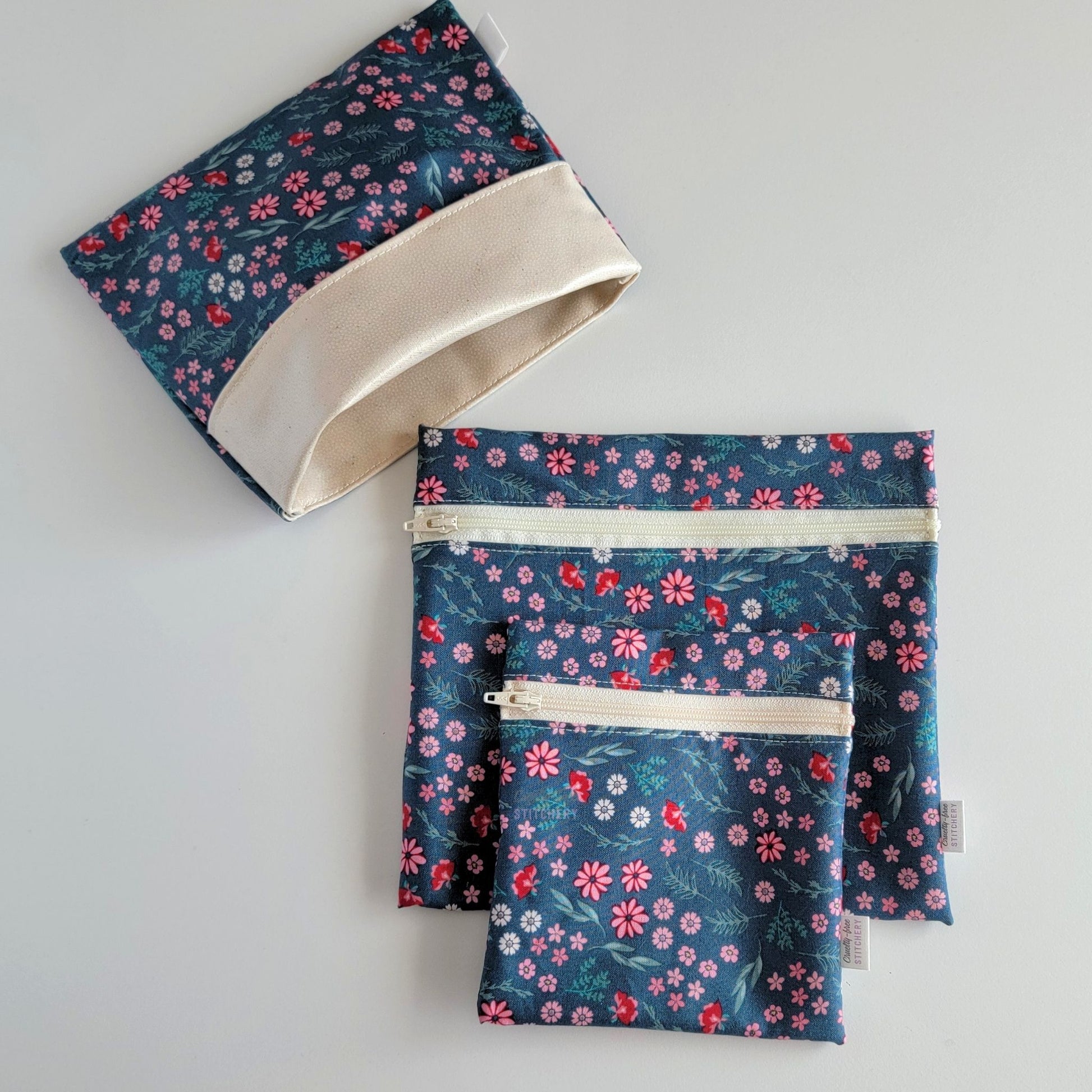 Set of three reusable snack bags with a deep teal and pink flowers print. Two have zippers, one large and one small, and one is a fold-over style. The fold-over one is unfolded to show the beige lining that matches the zippers. The print is a deep teal blue-green with small pink daisies and other flowers with greenery stems.
