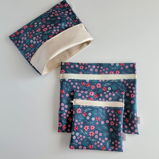 Set of three reusable snack bags with a deep teal and pink flowers print. Two have zippers, one large and one small, and one is a fold-over style. The fold-over one is unfolded to show the beige lining that matches the zippers. The print is a deep teal blue-green with small pink daisies and other flowers with greenery stems.
