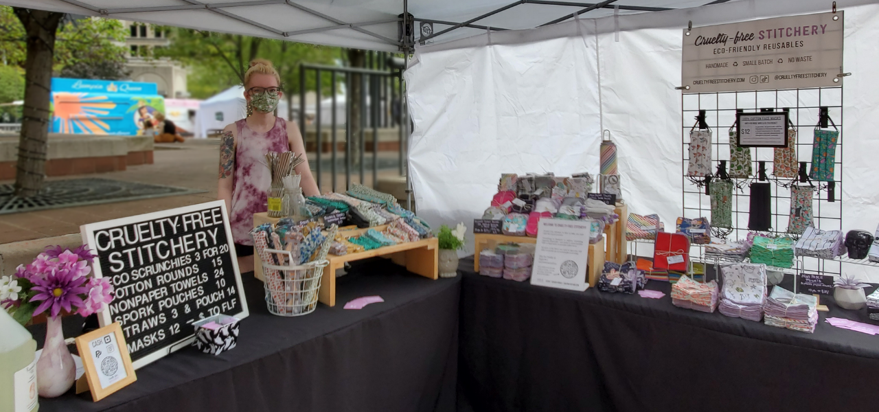 Panoramic photo of a Cruelty-Free Stitchery craft show booth. Outdoors under a white tent, with product set up on the tables. Abby is standing behind the table with a green mask and purple tie-dye tank top.