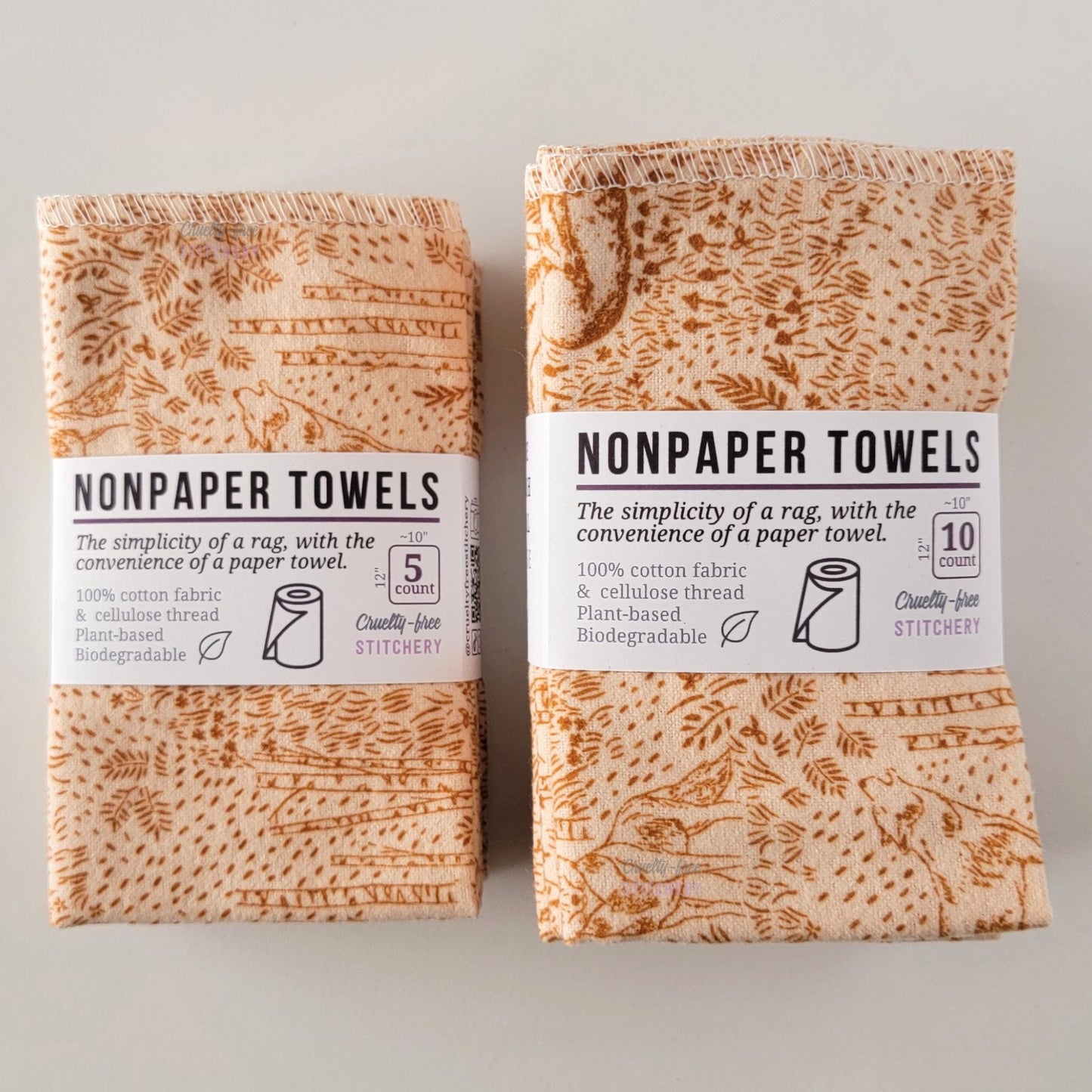 Two packages of the Acorn Woodland NonPaper Towels, one 5-pack and one 10-pack. Both wrapped with a paper tag, The tag also has the fiber content, a graphic of a paper towel roll, the Cruelty-Free Stitchery logo, and a rectangle with rounded corners that has the dimensions on the outside, 10x12", and the number of towels inside.