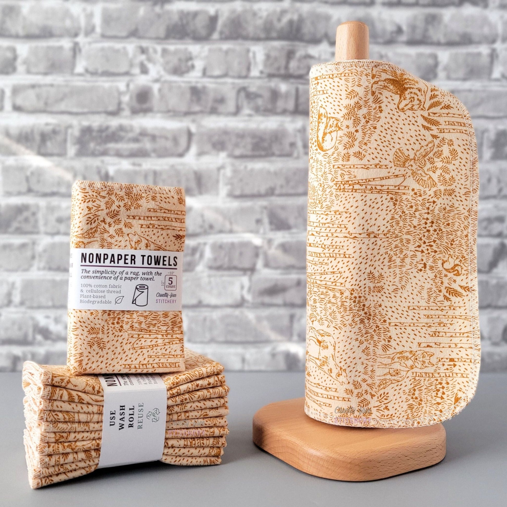 Acorn Woodland NonPaper Towels, displayed on a wooden paper towel holder next to packaged bundles. The print is a simple monochrome sketch in a burnt orange tan color, on a lighter tan background. There are all kinds of woodland creatures, trees, and other plants. Owls, wolves, foxes, squirrels, and bears.