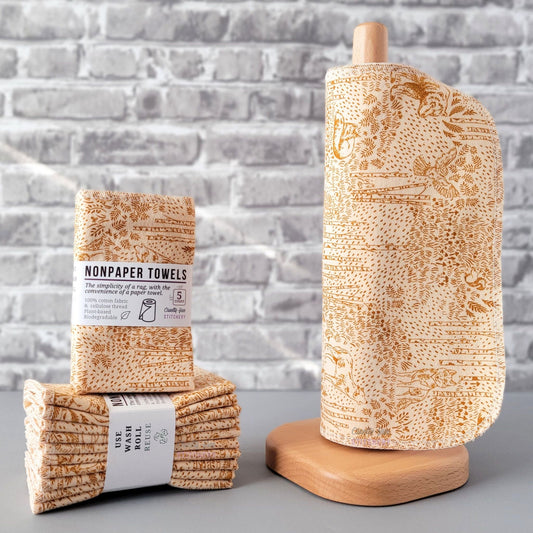 Acorn Woodland NonPaper Towels, displayed on a wooden paper towel holder next to packaged bundles. The print is a simple monochrome sketch in a burnt orange tan color, on a lighter tan background. There are all kinds of woodland creatures, trees, and other plants. Owls, wolves, foxes, squirrels, and bears.