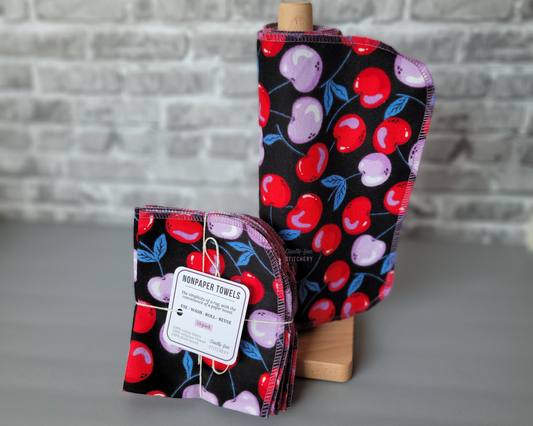 A set of black non-paper towels with a bright red and light purple cherries print, displayed on a wooden stand with a label showing the product name and size.