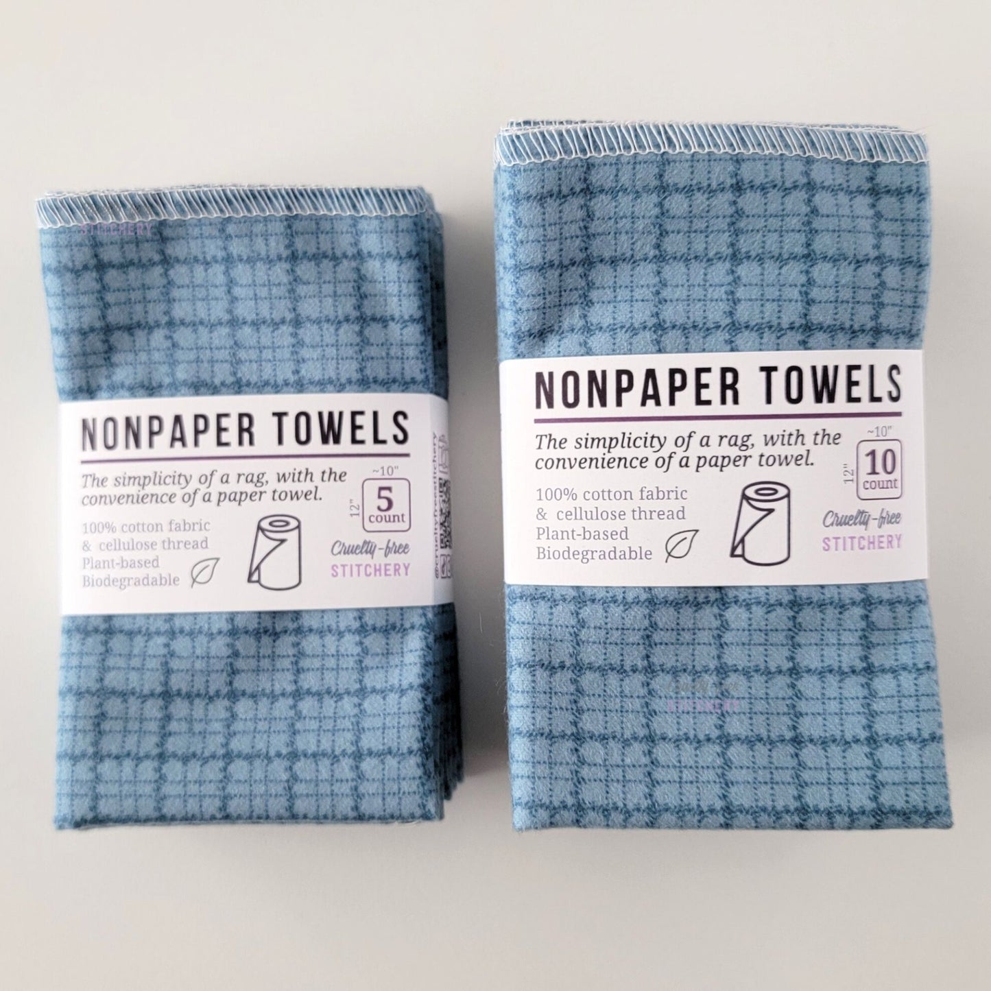 Two packages of the Blue Kitchen Plaid NonPaper Towels, one 5-pack and one 10-pack. Both wrapped with a paper tag, The tag also has the fiber content, a graphic of a paper towel roll, the Cruelty-Free Stitchery logo, and a rectangle with rounded corners that has the dimensions on the outside, 10x12", and the number of towels inside.