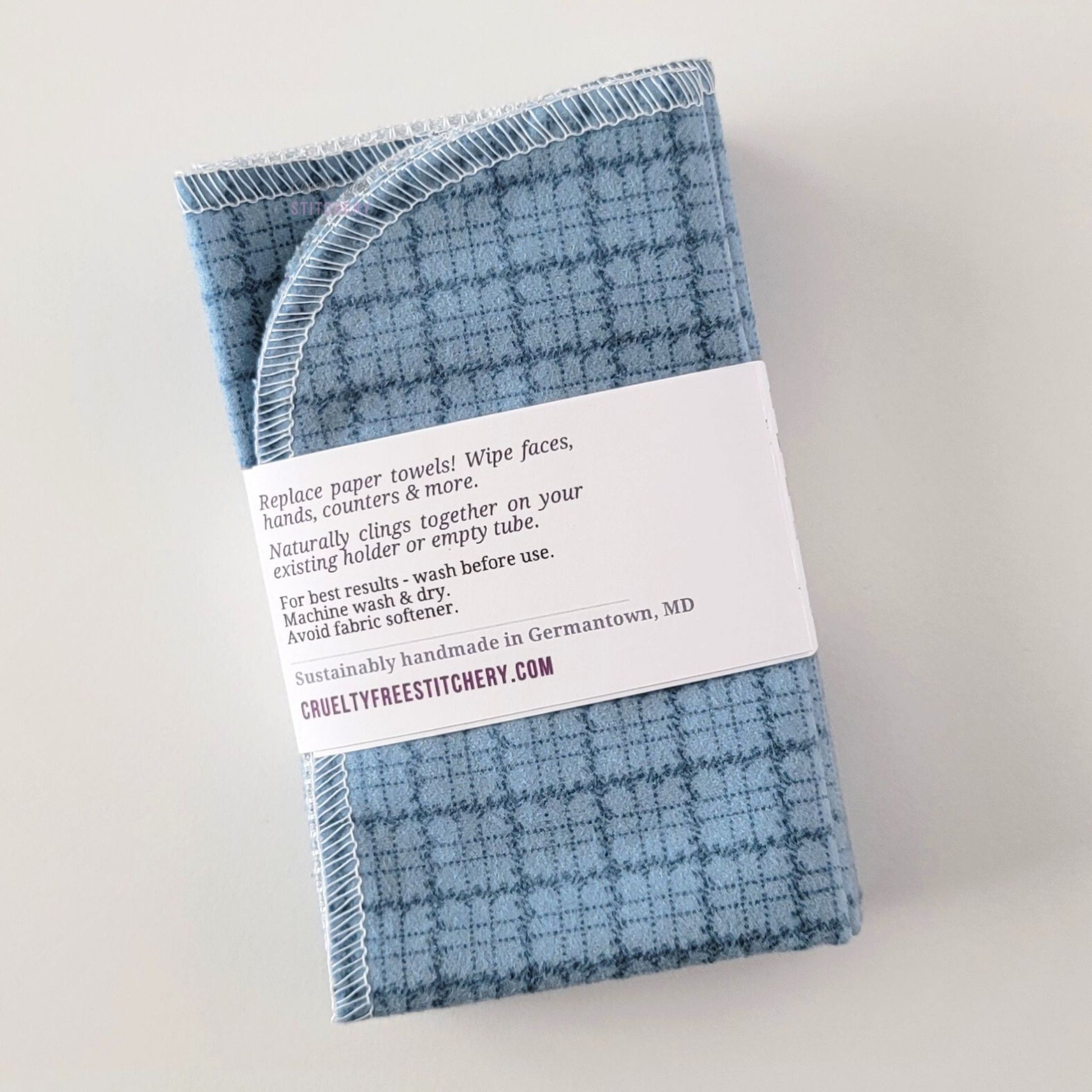 The back of a packaged bundle of the Blue Kitchen Plaid NonPaper Towels. The tag reads "Replace paper towels! Wipe faces, hands, counters, and more. Naturally clings together on your existing holder or empty tube. For best results - wash before use. Machine wash and dry. Avoid fabric softener. Sustainably handmade in Germantown, Maryland. CrueltyFreeStitchery dot com."