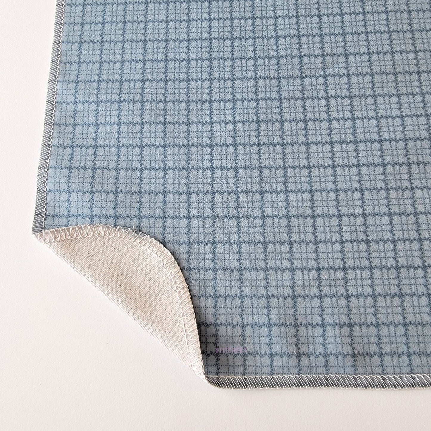 The Blue Kitchen Plaid NonPaper Towel, shown close-up with the bottom corner folded back to show they are unprinted on the back side, and are one layer.