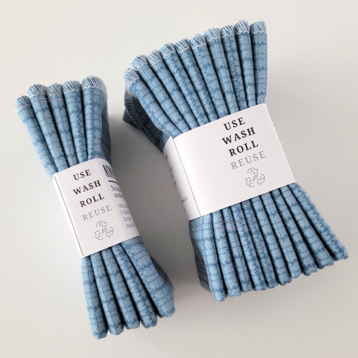 Two packages of the Blue Kitchen Plaid NonPaper Towels, shown on their side so display the 5-pack and 10-packs. The side of the paper tag says "use, wash, roll, reuse" and has the recycling logo with a leaf in the center, to symbolize they are biodegradable.
