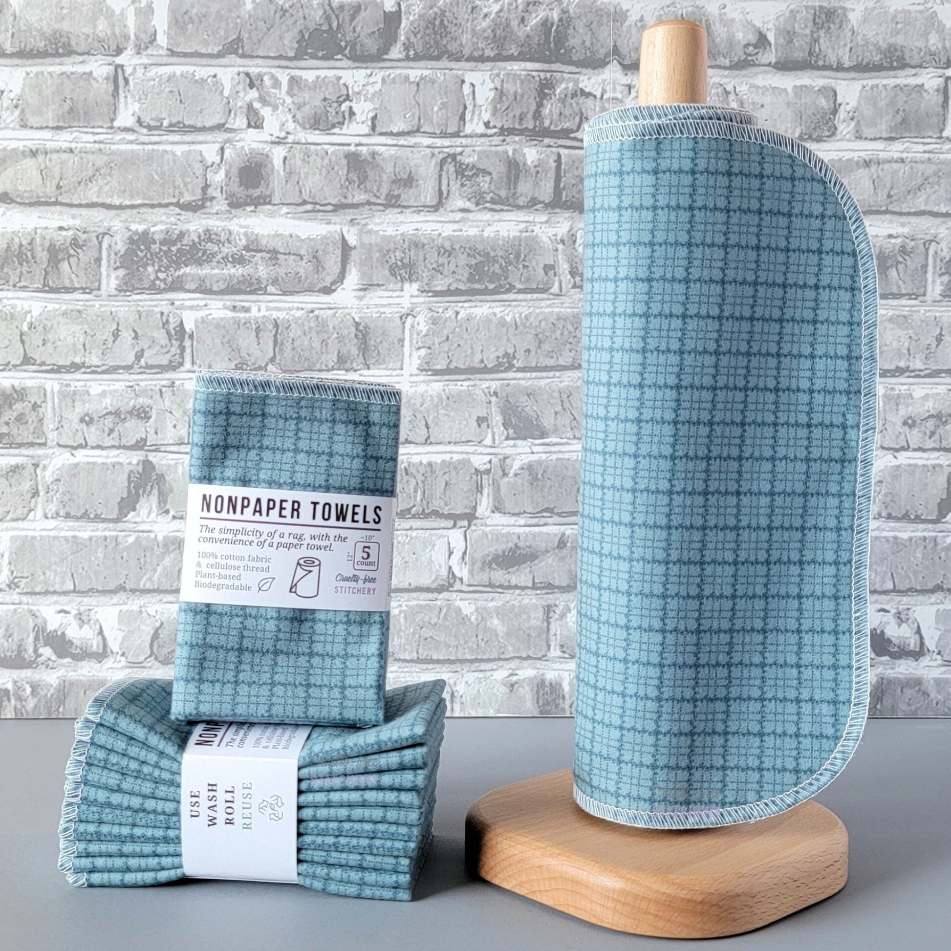 Blue Kitchen Plaid NonPaper Towels, displayed on a wooden paper towel holder next to packaged bundles. They are an sky blue with darker blue plaid lines. The lines are more scribbly when you look closer. The plaid squares are approximately half an inch.