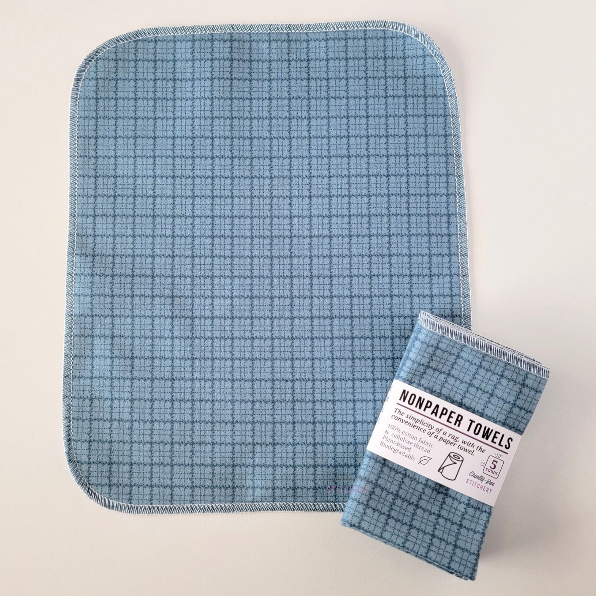 An unfolded Blue Kitchen Plaid NonPaper Towel, next to a bundled 5-pack. The towel is a rectangle with rounded corners, stitched around the edges with white thread. The bundled pack is folded up with a paper wrapper that says "NonPaper Towels, the simplicity of a rag with the convenience of a paper towel"