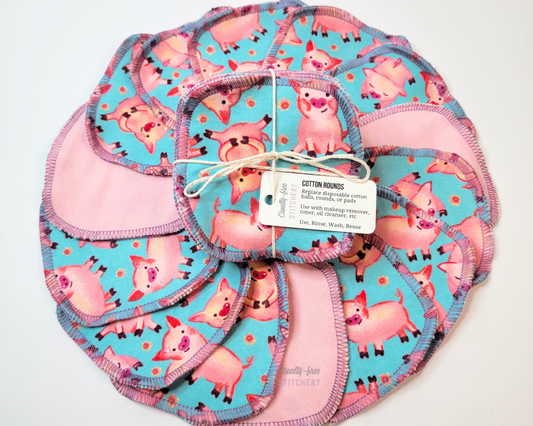 Blue with pigs cotton rounds. Bright aqua blue with cute pink pigs and tiny pink flowers. Pictured as a bundle on top of more rounds arranged in a circle, some flipped over to show the back is a solid light pink. Stitched in lilac purple.