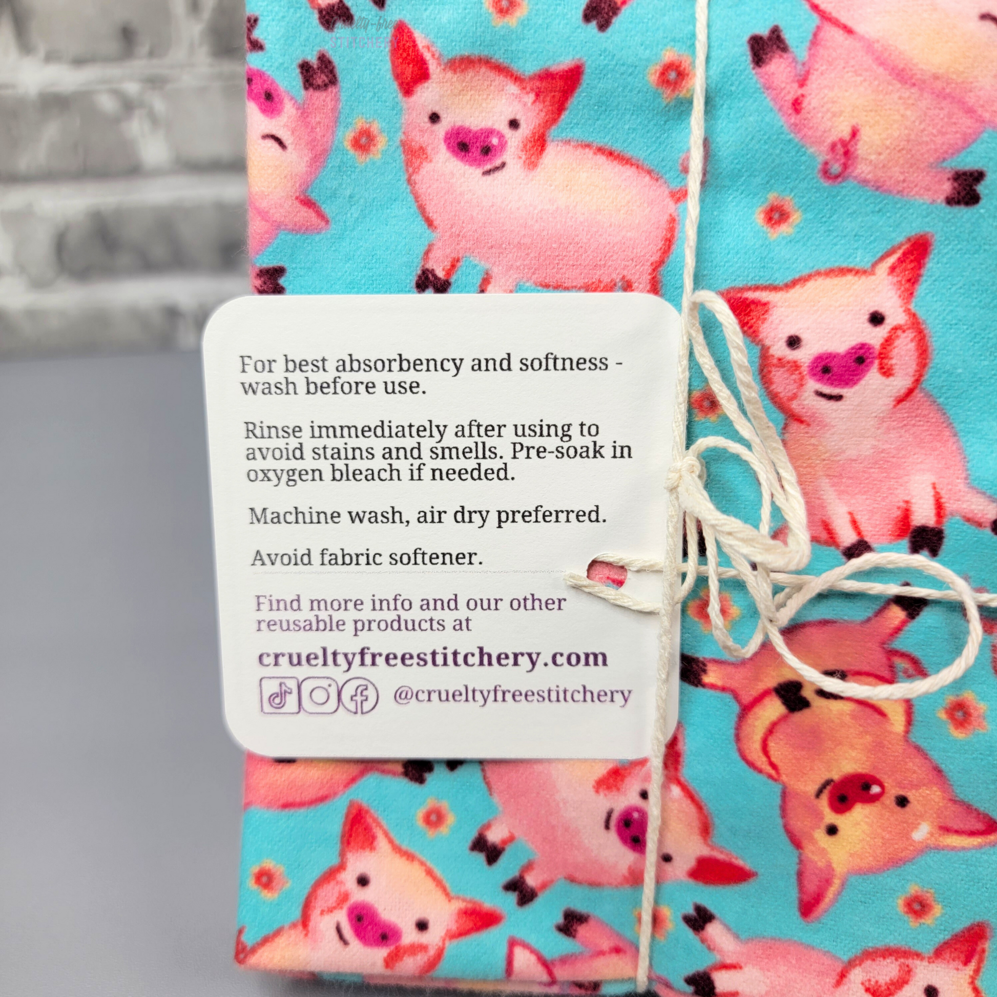 A close-up of the back of the tag on the blue pigs NonPaper Towels. It says "for best absorbency and softness, wash before use. Rinse immediately after using to avoid stains and smells. Pre-soak in oxygen bleach if needed. Machine wash, air dry preferred. Avoid fabric softener. Find more info and our other reusable products at CrueltyFreeStitchery dot com. tiktok, instagram, facebook at CrueltyFreeStitchery