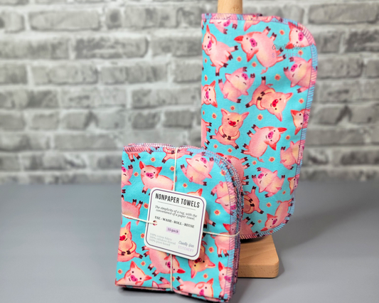 Aqua Blue with pink pigs print NonPaper Towels. They are rolled onto a wooden paper towel holder, and sitting next to a bundled pack of 10. The bundled pack is folded and tied with a string, the tag says "nonpaper towels, use, wash, roll, reuse, the simplicity of a rag with the convenience of a paper towel." The edges of each NonPaper Towel are stitched with a lilac purple thread, with the corners rounded.