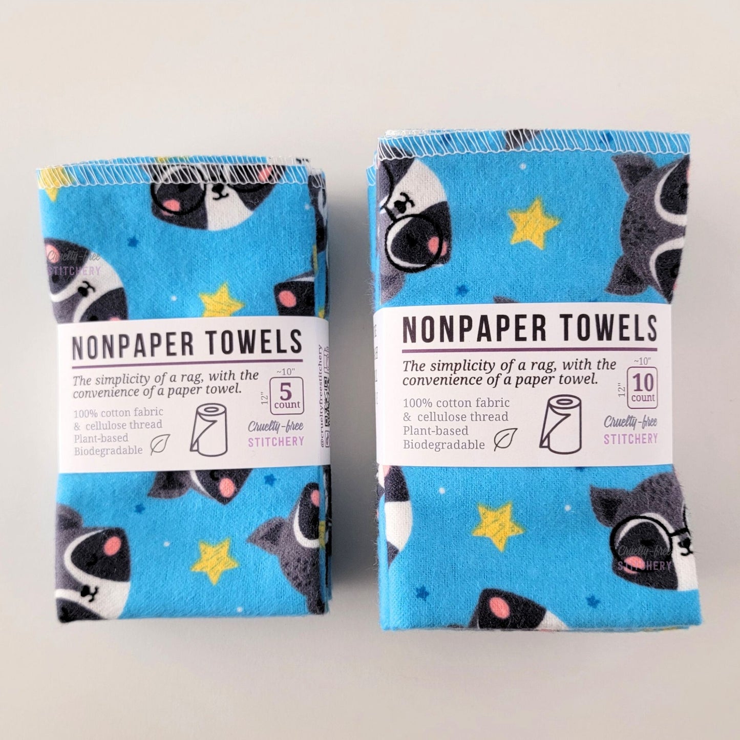 Two packages of the Blue Raccoons NonPaper Towels, one 5-pack and one 10-pack. Both wrapped with a paper tag, The tag also has the fiber content, a graphic of a paper towel roll, the Cruelty-Free Stitchery logo, and a rectangle with rounded corners that has the dimensions on the outside, 10x12", and the number of towels inside.