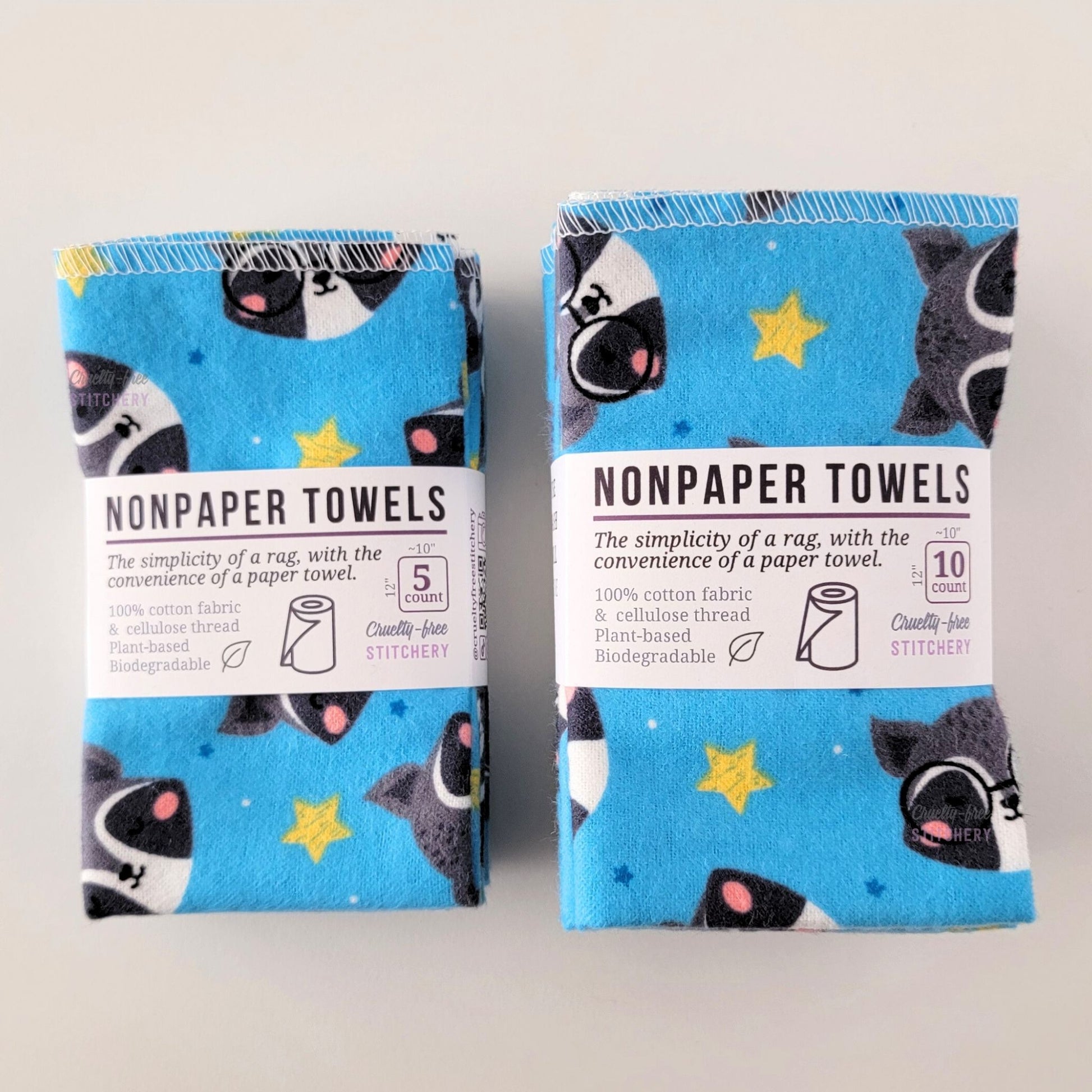 Two packages of the Blue Raccoons NonPaper Towels, one 5-pack and one 10-pack. Both wrapped with a paper tag, The tag also has the fiber content, a graphic of a paper towel roll, the Cruelty-Free Stitchery logo, and a rectangle with rounded corners that has the dimensions on the outside, 10x12", and the number of towels inside.