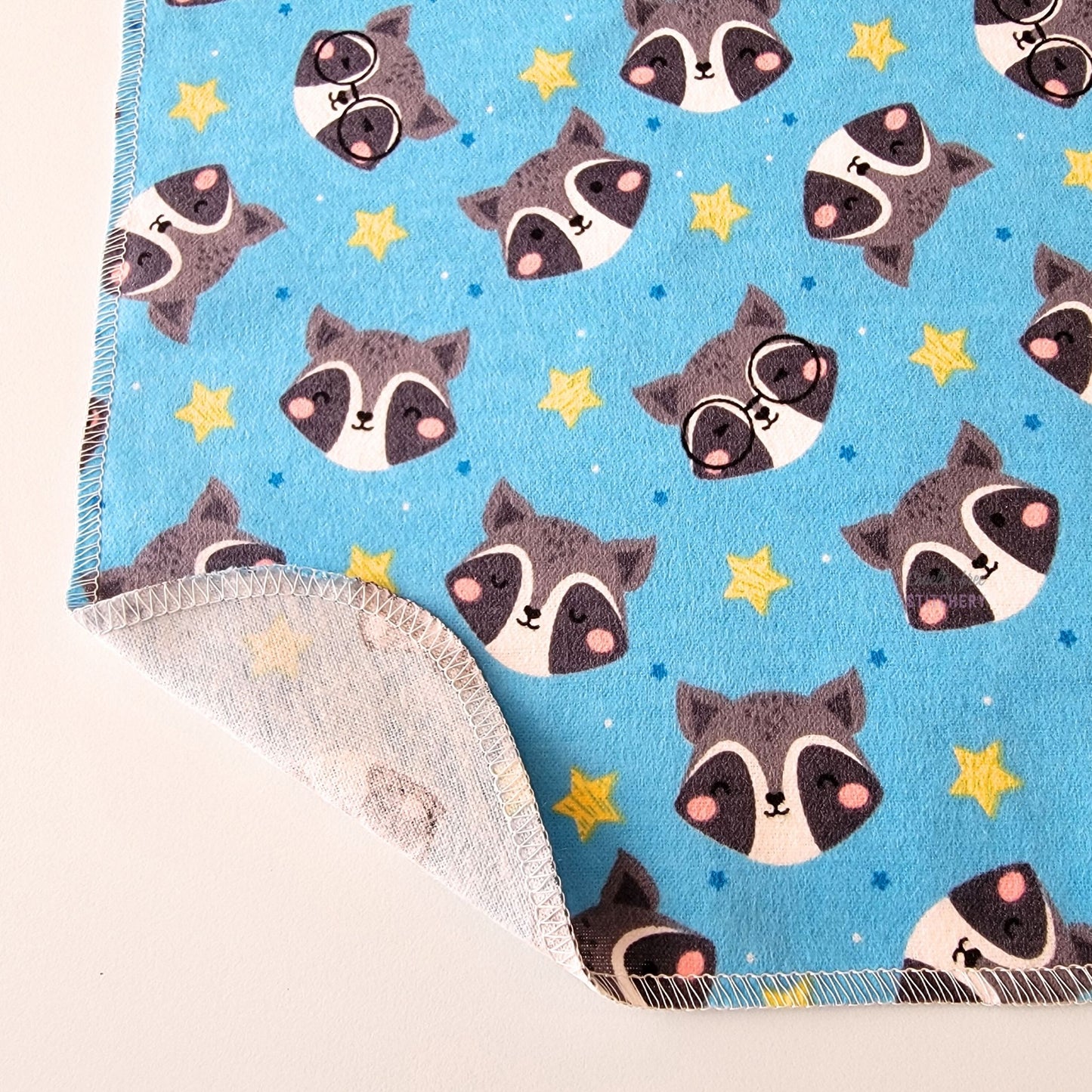 The Blue Raccoons NonPaper Towel, shown close-up with the bottom corner folded back to show they are unprinted on the back side, and are one layer.