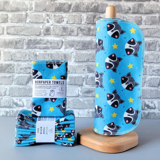 Blue with raccoon faces NonPaper Towels, displayed on a wooden paper towel holder next to packaged bundles. The print is a bright sky blue with cute raccoon faces, some of them have glasses, and there are yellow stars and smaller blue stars scattered around. 