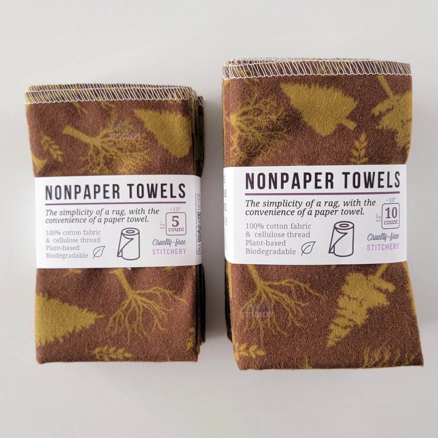 Two packages of the Brown Trees NonPaper Towels, one 5-pack and one 10-pack. Both wrapped with a paper tag, The tag also has the fiber content, a graphic of a paper towel roll, the Cruelty-Free Stitchery logo, and a rectangle with rounded corners that has the dimensions on the outside, 10x12", and the number of towels inside.