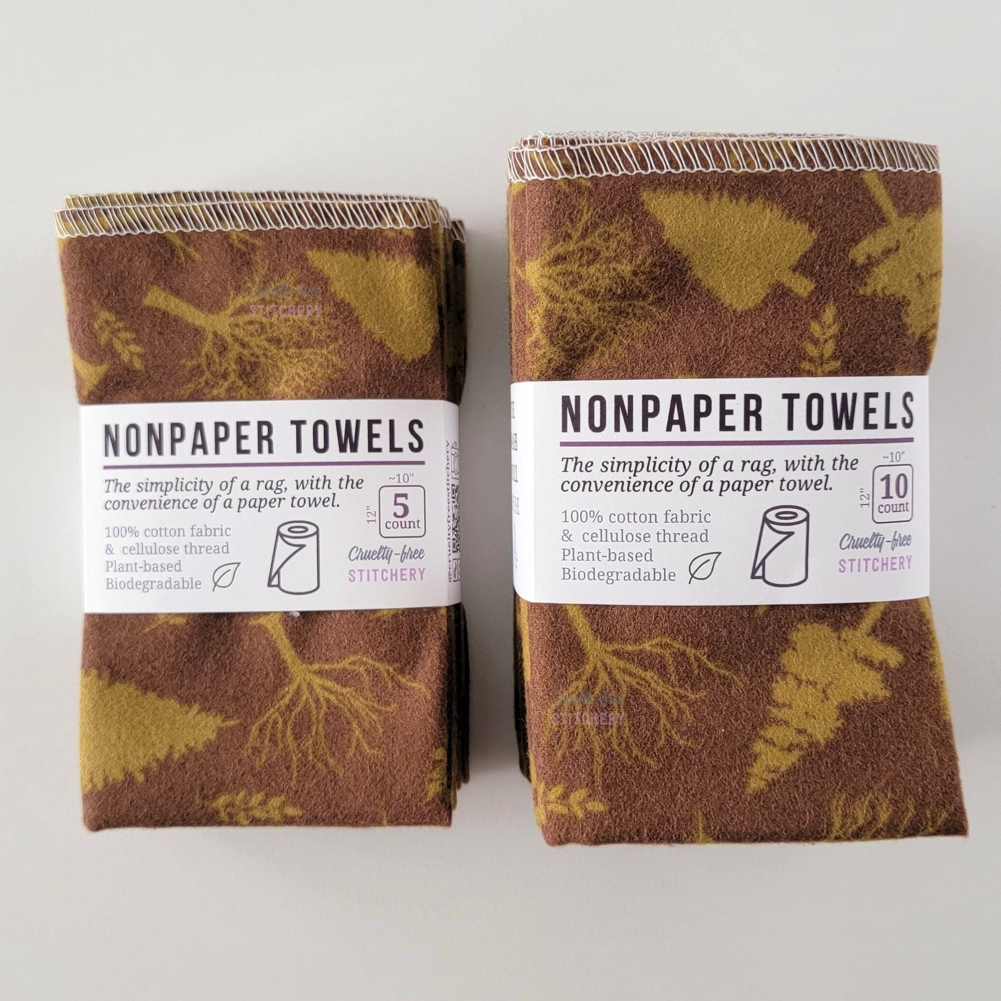 Two packages of the Brown Trees NonPaper Towels, one 5-pack and one 10-pack. Both wrapped with a paper tag, The tag also has the fiber content, a graphic of a paper towel roll, the Cruelty-Free Stitchery logo, and a rectangle with rounded corners that has the dimensions on the outside, 10x12", and the number of towels inside.