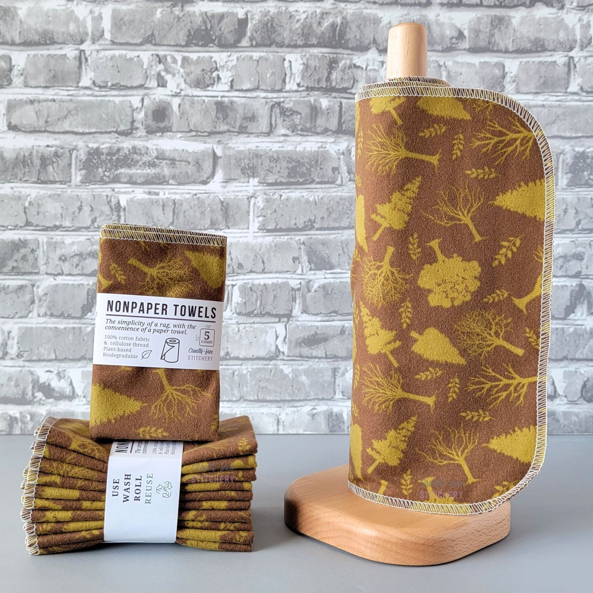 Brown with Green Trees NonPaper Towels, displayed on a wooden paper towel holder next to packaged bundles. The print is a chocolate brown background with various kinds of trees in a solid light leaf green color. The trees are scattered around and a little over an inch tall.
