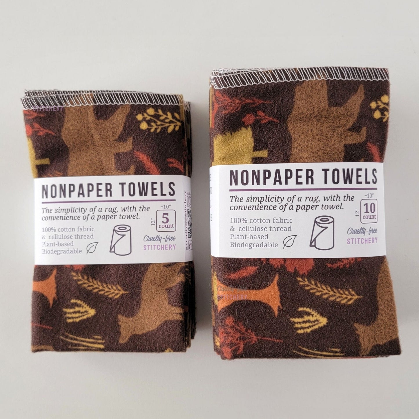 Two packages of the Brown Woodland NonPaper Towels, one 5-pack and one 10-pack. Both wrapped with a paper tag, The tag also has the fiber content, a graphic of a paper towel roll, the Cruelty-Free Stitchery logo, and a rectangle with rounded corners that has the dimensions on the outside, 10x12", and the number of towels inside.