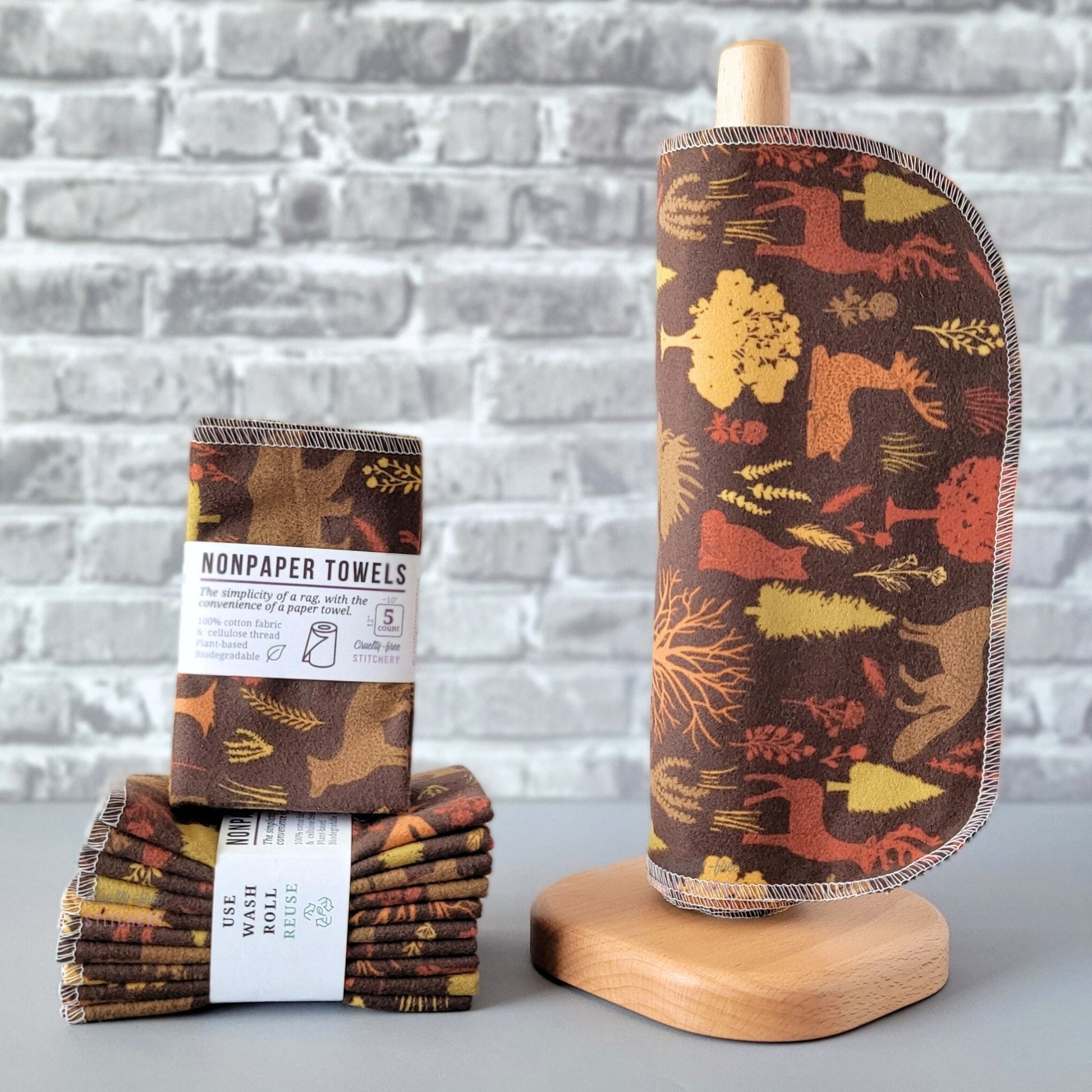 Brown Woodland NonPaper Towels, displayed on a wooden paper towel holder next to packaged bundles. The print is a dark brown with silhouettes of various trees, flowers, and woodland creatures, in various shades of tan, yellow, light orange, and burnt orange. Animals are deer, foxes, bears, and moose.