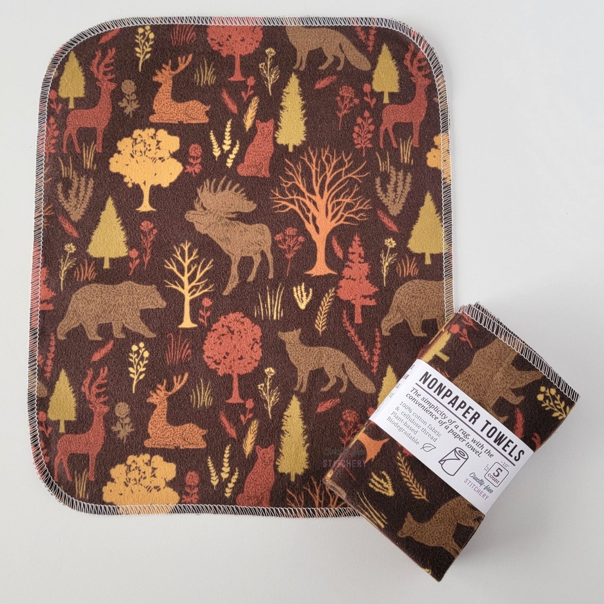 An unfolded Brown Woodland NonPaper Towel, next to a bundled 5-pack. The towel is a rectangle with rounded corners, stitched around the edges with white thread. The bundled pack is folded up with a paper wrapper that says "NonPaper Towels, the simplicity of a rag with the convenience of a paper towel"
