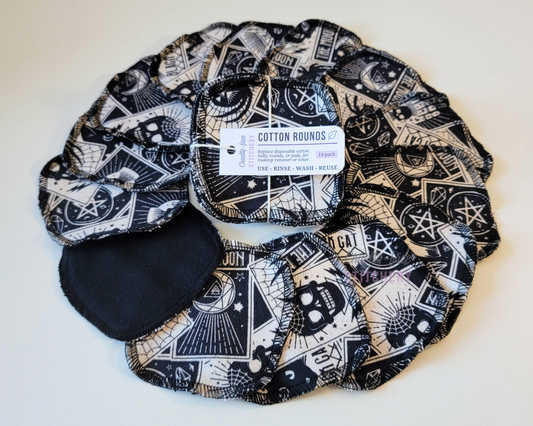 Reusable Cotton Rounds with a scattered Tarot card print on the front, solid black on the back. The print is black and off-white, various skulls, cats, pentagrams, moons, etc. They are a rounded square shape and stitched around the edges with black thread. This photo is a bundled pack that is tied with string and a small paper tag, sitting in the center of more cotton rounds arranged in a circle.