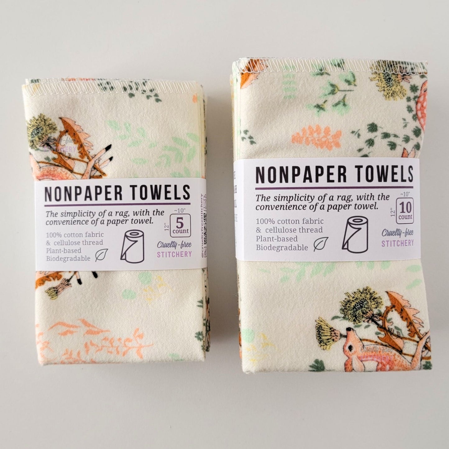 Two packages of the Dandelion Doe NonPaper Towels, one 5-pack and one 10-pack. Both wrapped with a paper tag, The tag also has the fiber content, a graphic of a paper towel roll, the Cruelty-Free Stitchery logo, and a rectangle with rounded corners that has the dimensions on the outside, 10x12", and the number of towels inside.